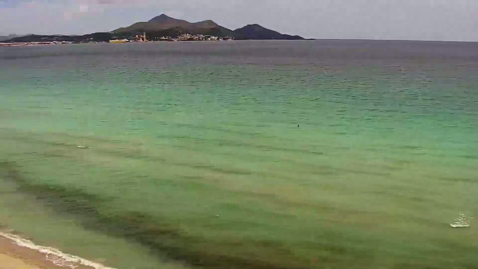 A tranquil beach scene shows calm, light-green water lapping gently at a sandy shore, with a distant mountain range visible under a mostly cloudy sky.