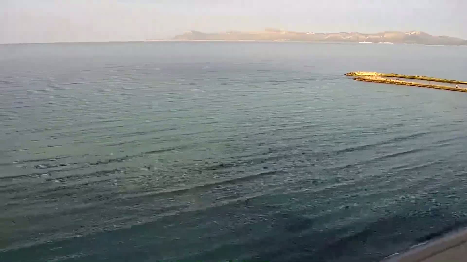 A calm, somewhat hazy day shows a long, thin breakwater extending into a body of water with a distant landmass visible on the horizon.