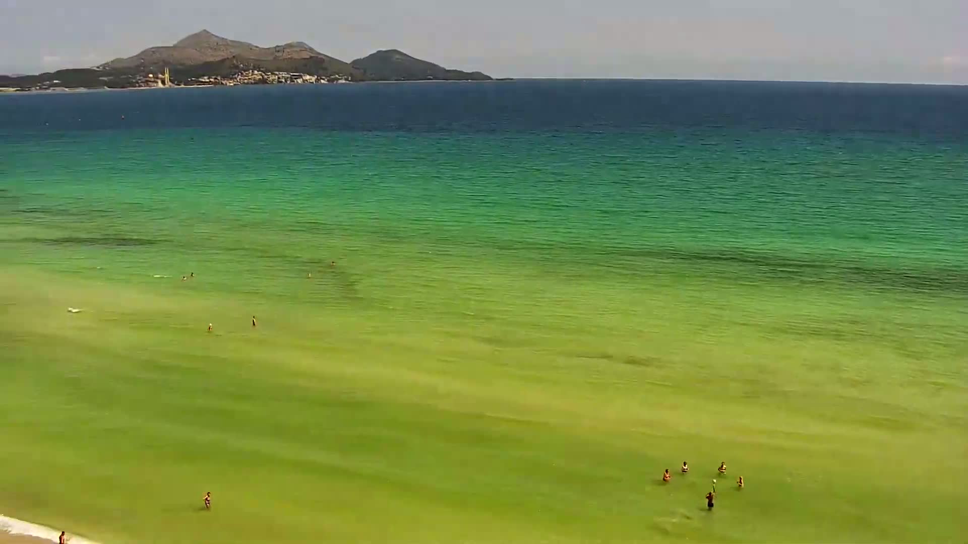 An aerial view shows a shallow, green-tinged ocean with several people swimming near a beach on a partly sunny day, with a distant mountain range visible on the horizon.