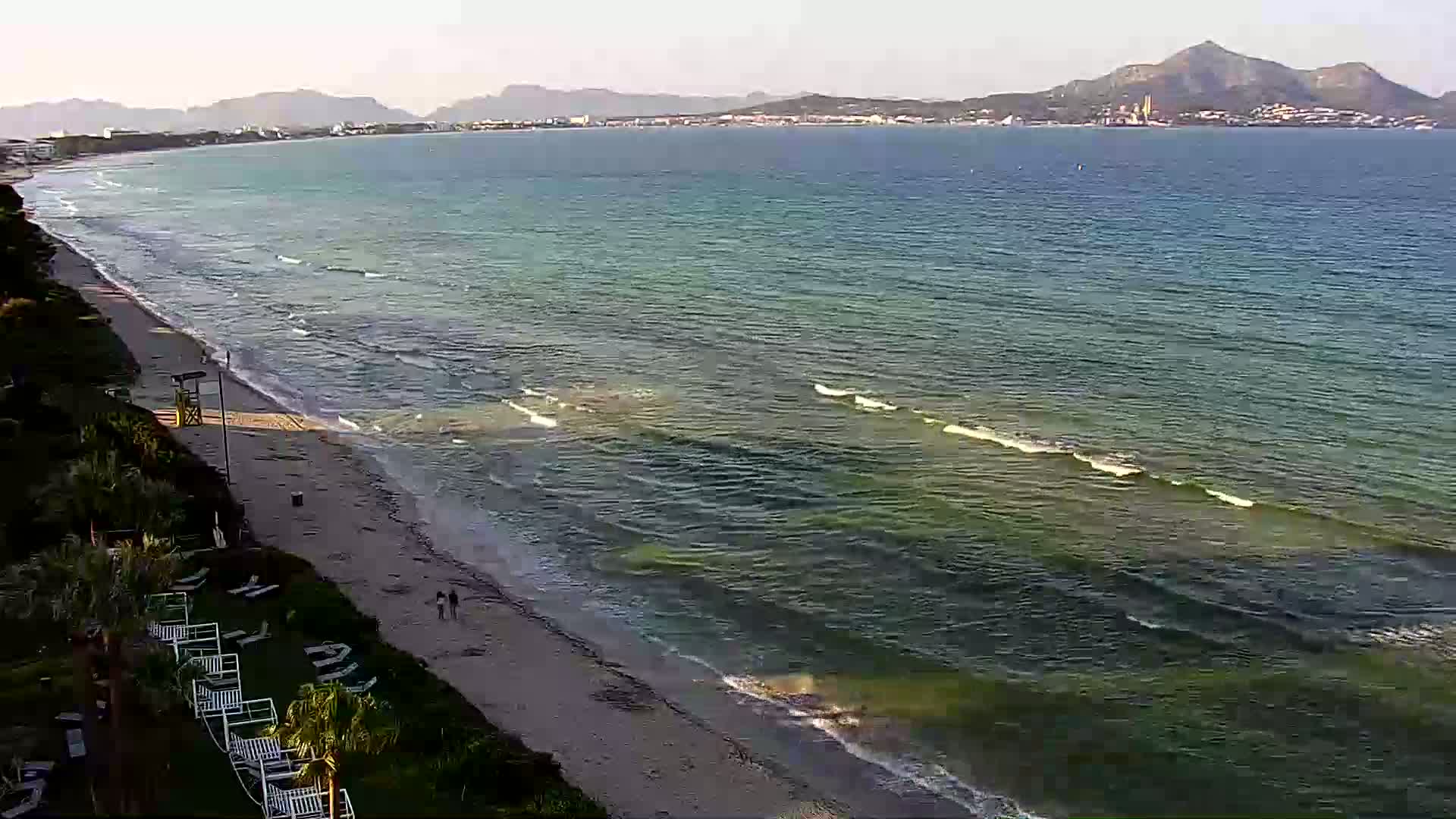 A sandy beach with calm, greenish-blue water and small waves laps the shore under a clear sky with distant mountains visible on the horizon.