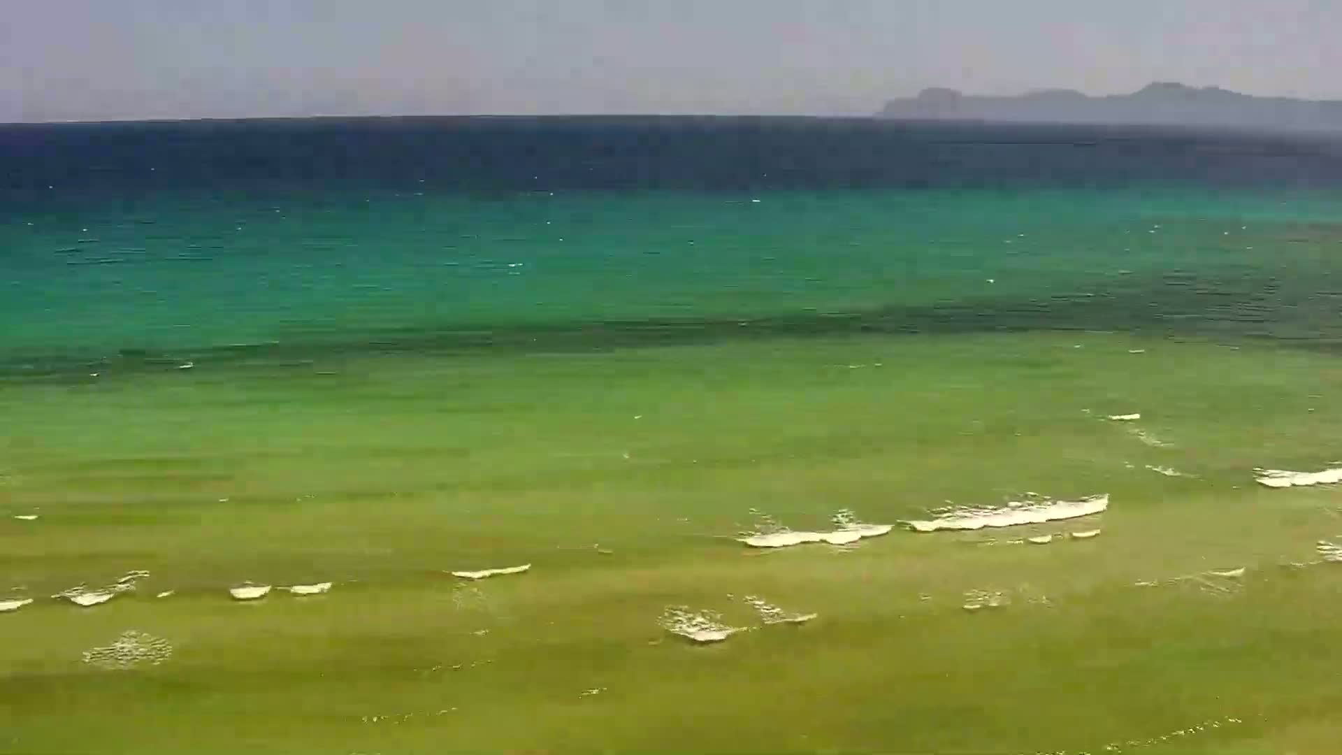 A calm sea with varying shades of green and blue water laps gently against a shallow, green shoreline under a hazy sky.