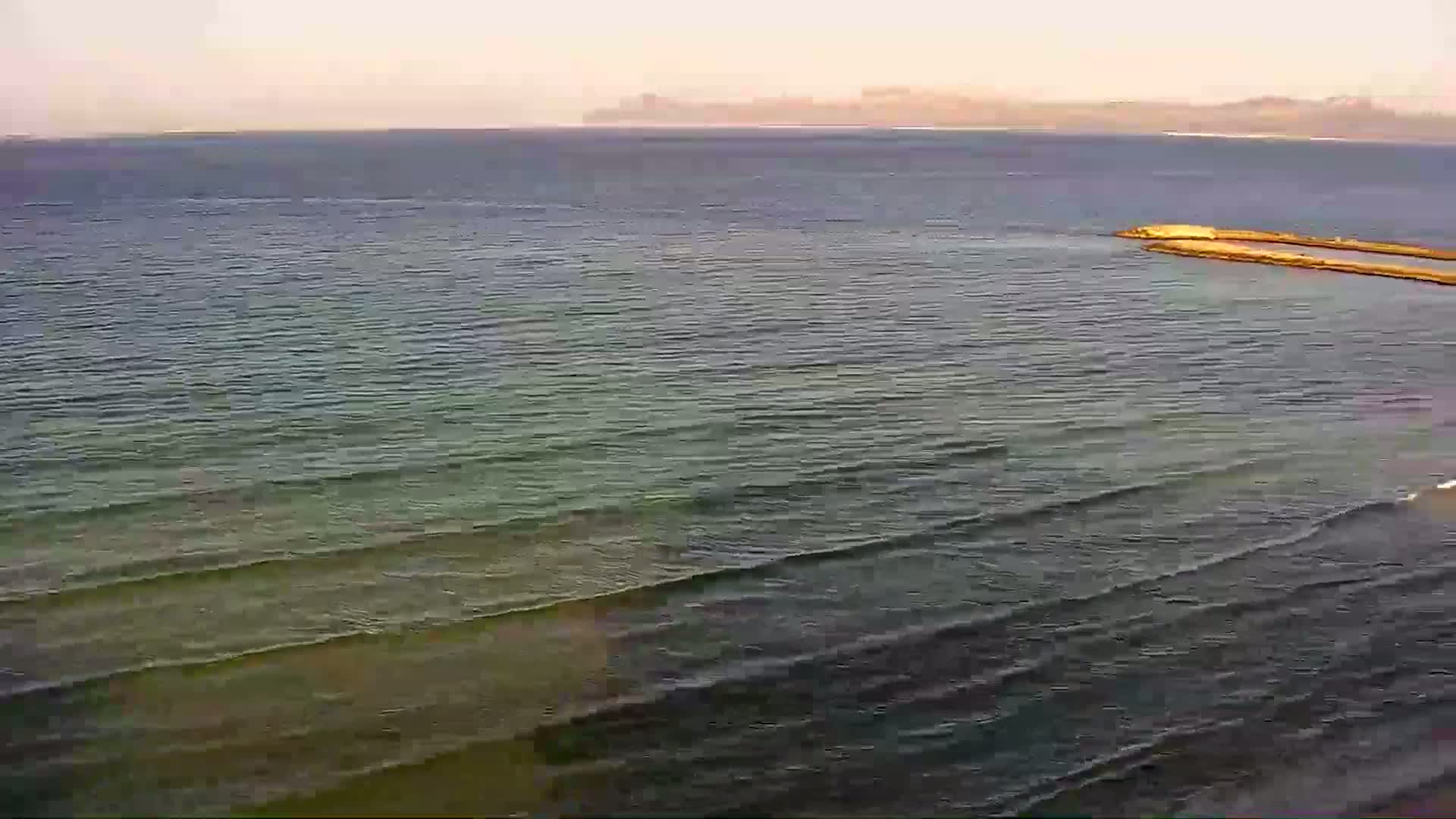 A calm sea with small waves laps gently against a man-made jetty under a hazy sky.