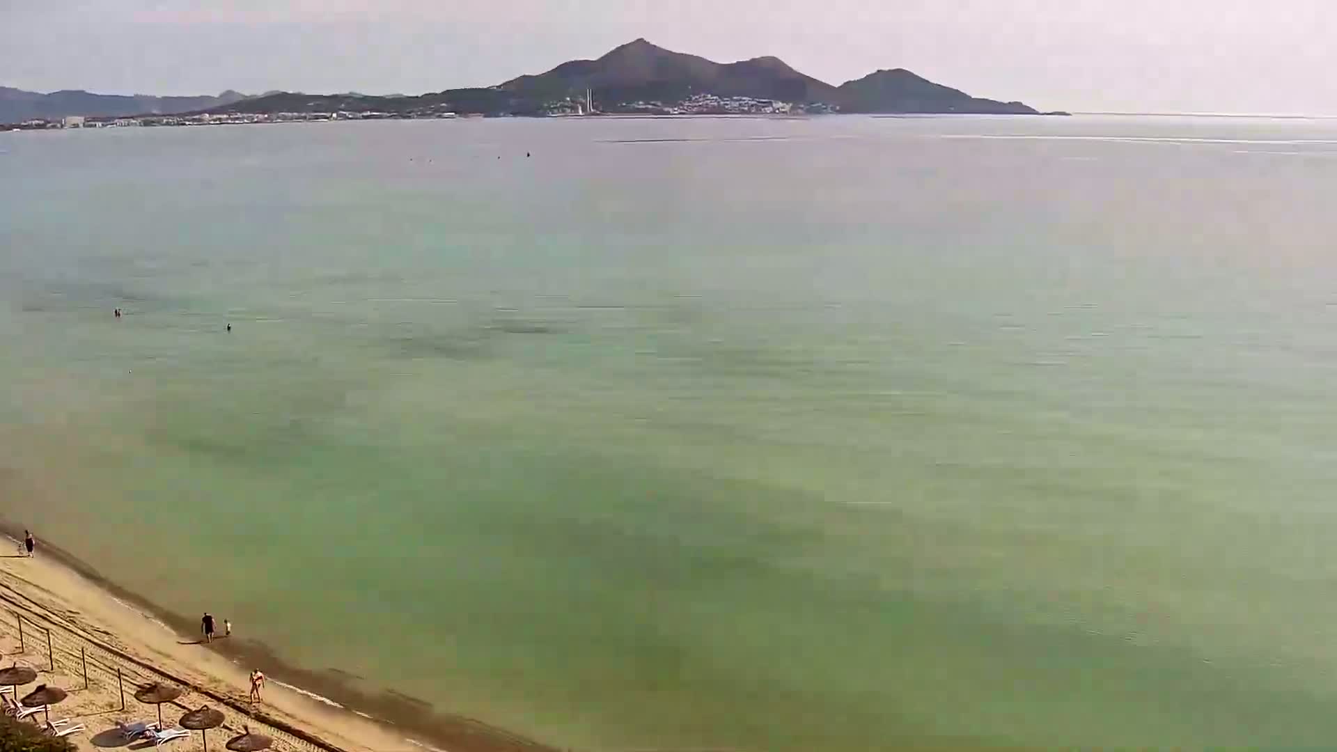 A sandy beach with a few people on it slopes down to a calm, light green sea with a distant mountain range visible on the horizon under a mostly clear sky.