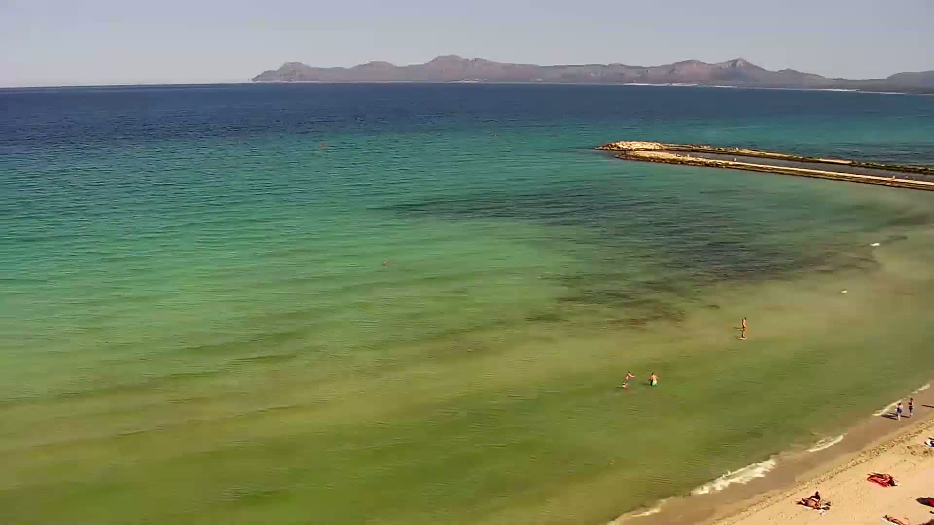 A sandy beach with calm, shallow, green-tinged water and a few people swimming and sunbathing, with a distant mountain range visible under a clear sky.