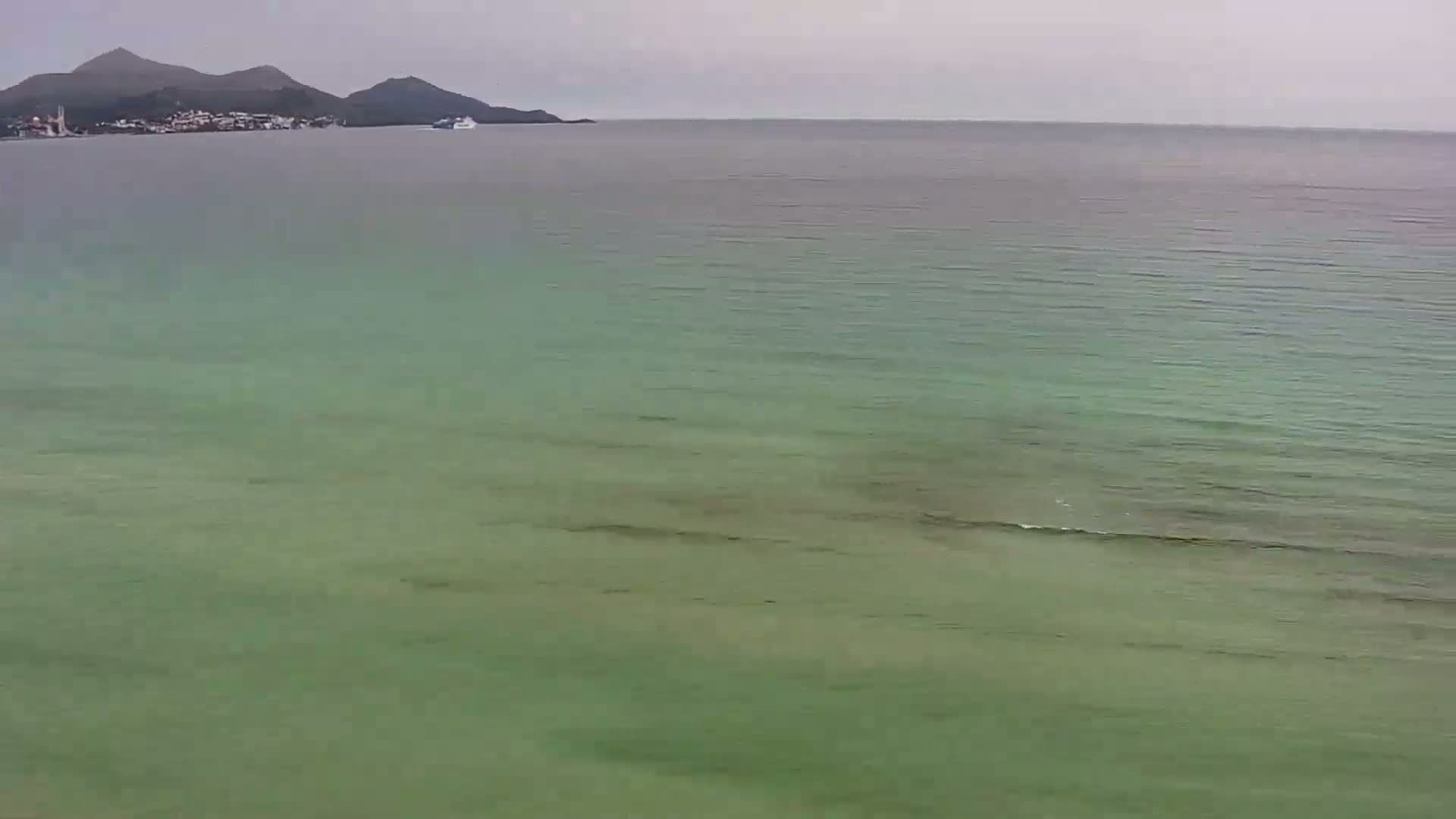 A calm, green-tinged sea stretches to a distant shoreline featuring dark mountains under an overcast sky.