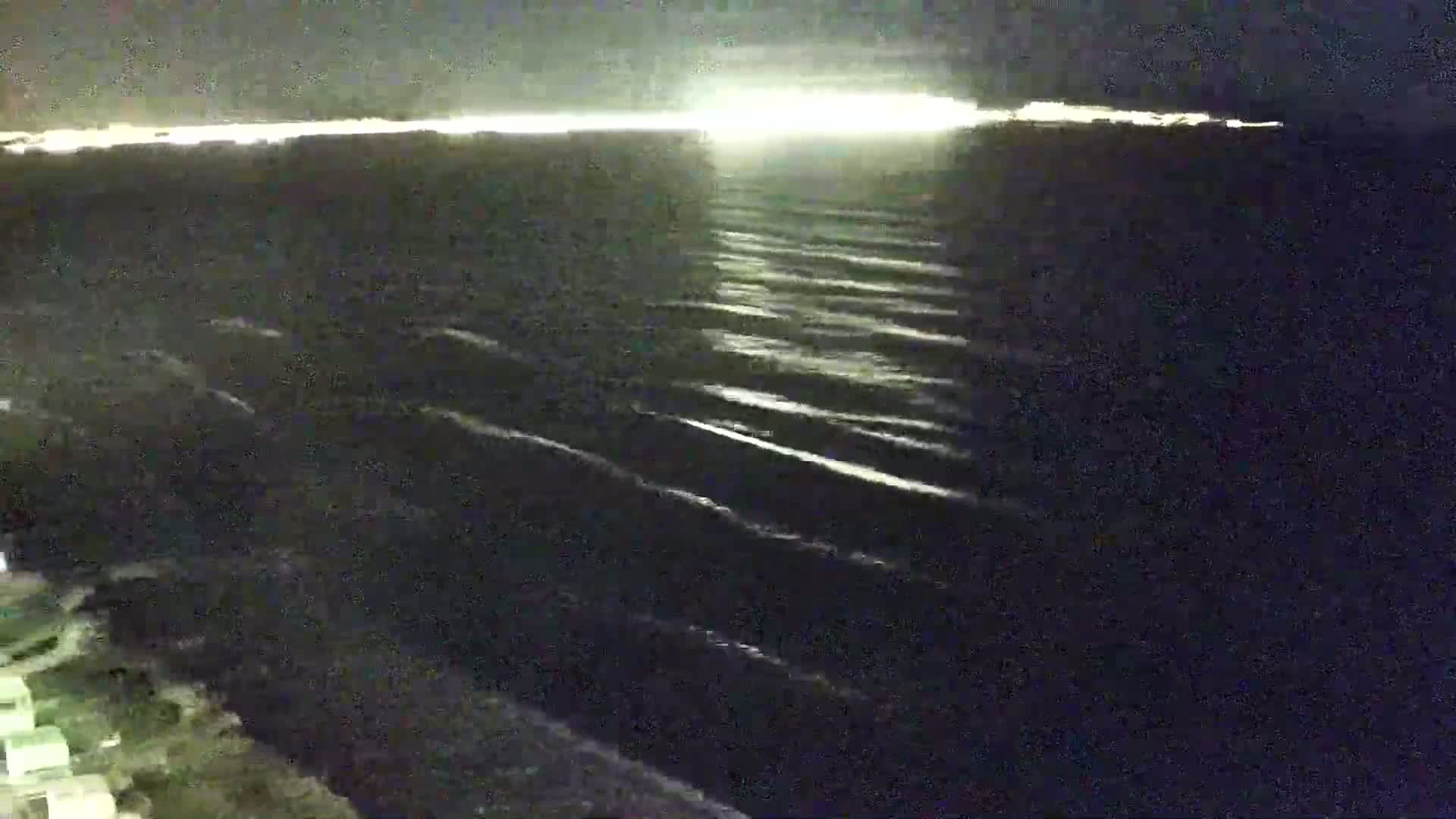 A dark, nighttime view of gently rippling water with a brightly lit shoreline in the distance.