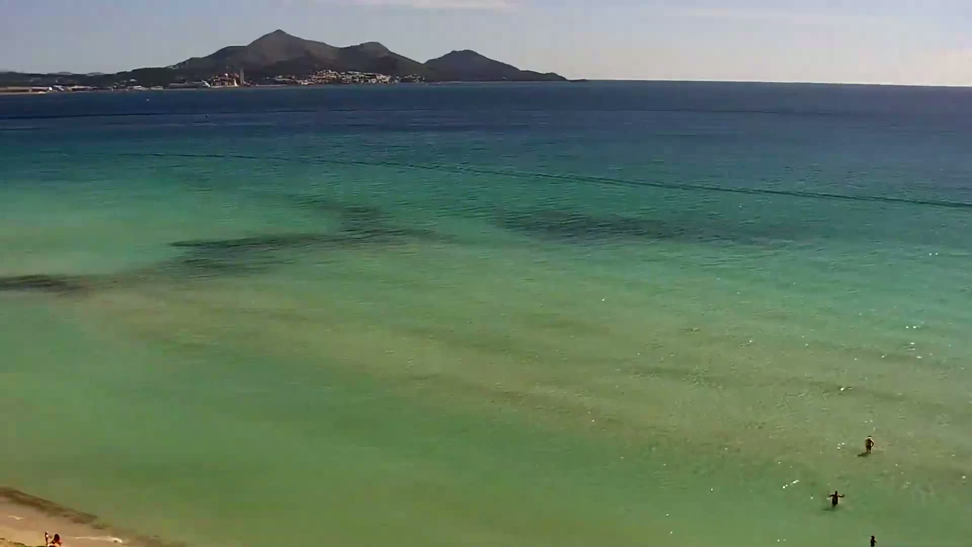 A tranquil, shallow, green-tinged ocean stretches to a distant mountainous coastline under a sunny sky.