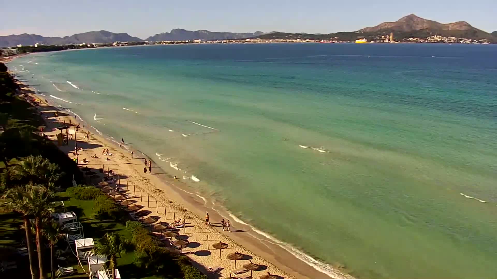 An aerial view shows a sandy beach with scattered beach umbrellas and people enjoying a sunny day with calm, shallow, clear turquoise water and distant mountains visible across the bay.