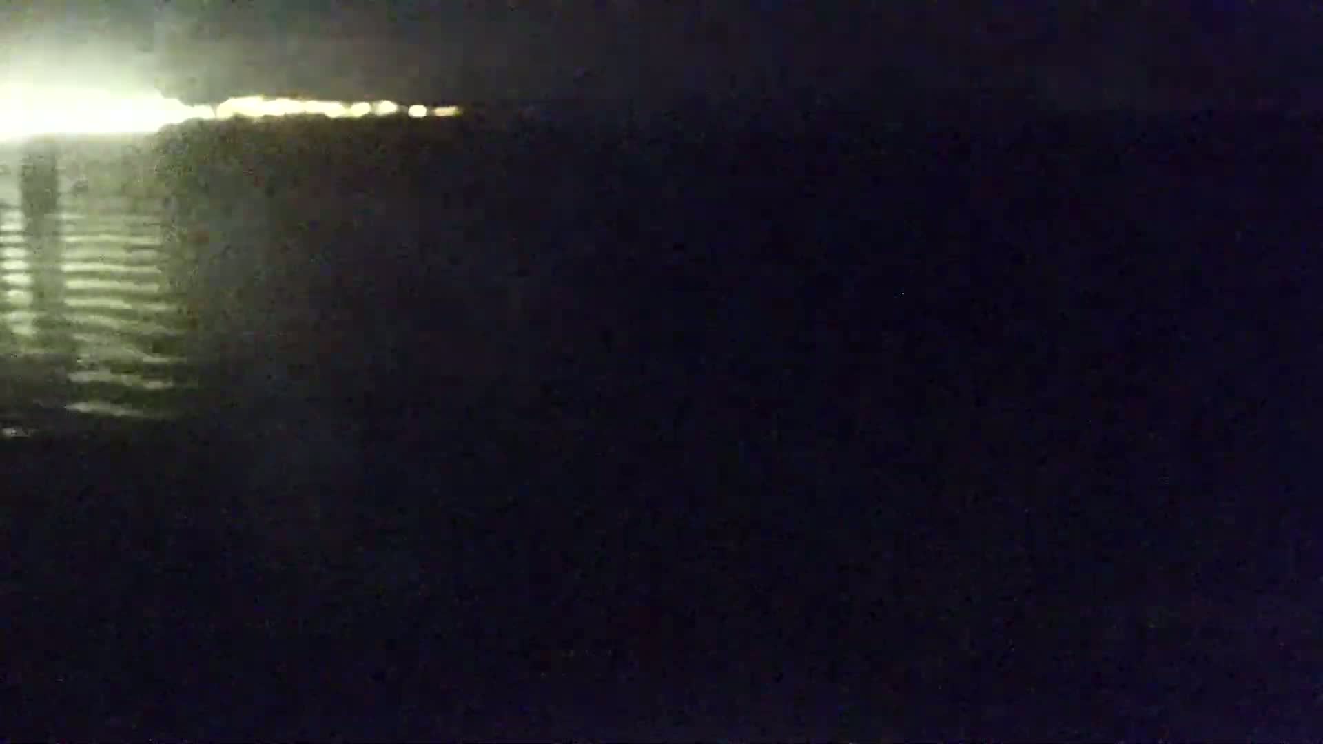 At night, a blurry view shows dark water reflecting a line of distant lights.
