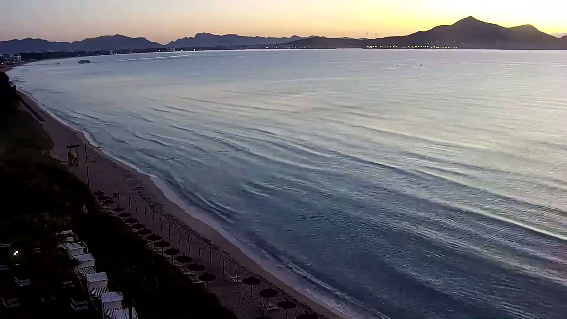A sandy beach with rows of closed umbrellas overlooks a calm sea at dawn, with mountains visible in the distance.