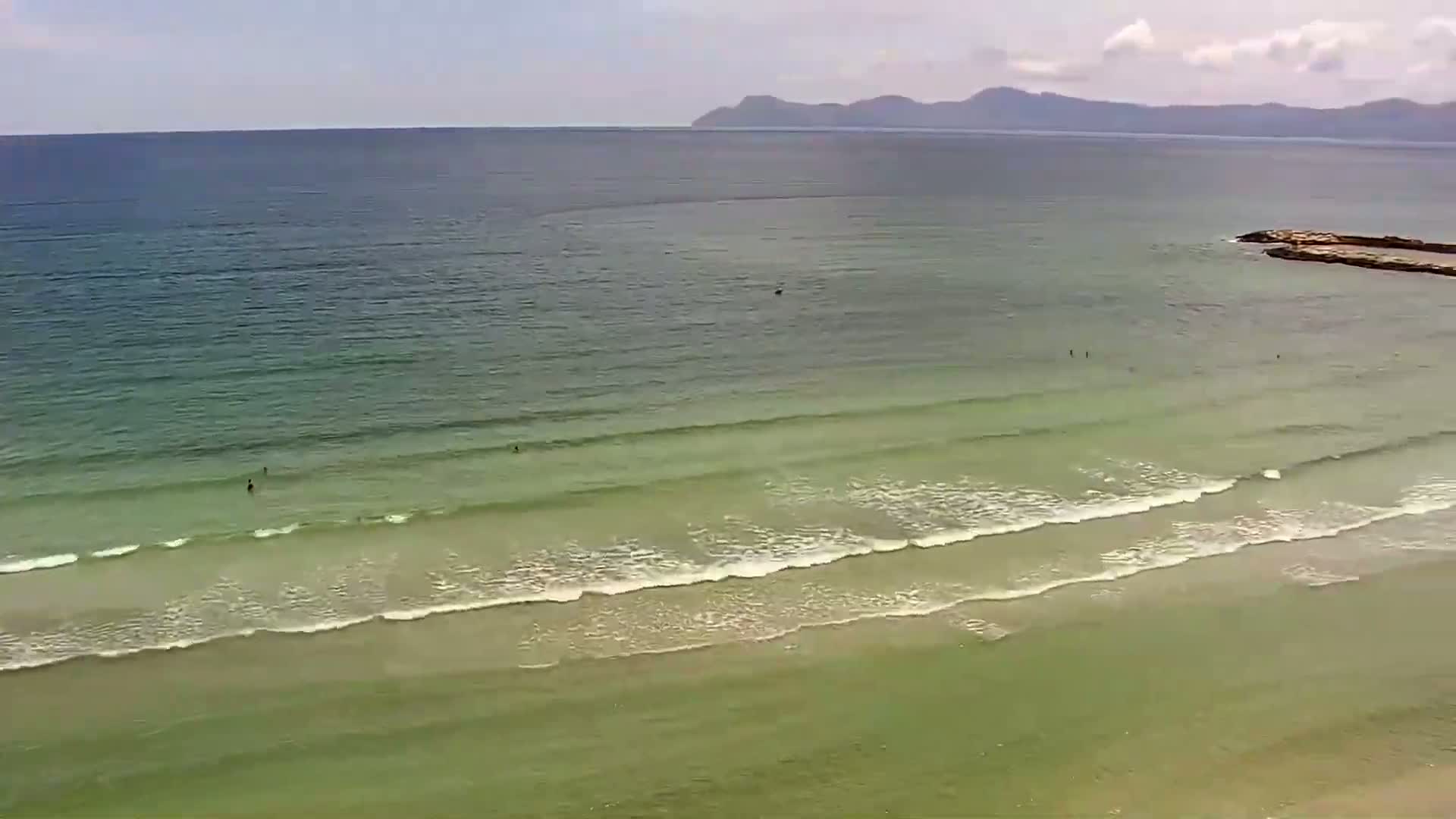A beach with light green water and small waves laps a sandy shore under a partly cloudy sky, with distant mountains visible on the horizon.