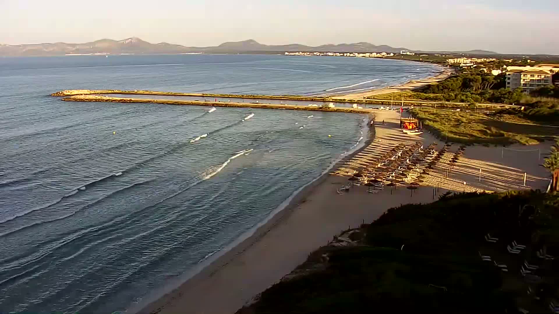 A sandy beach with calm waves and numerous beach umbrellas is situated next to a calm ocean under a clear sky, with distant mountains visible on the horizon.