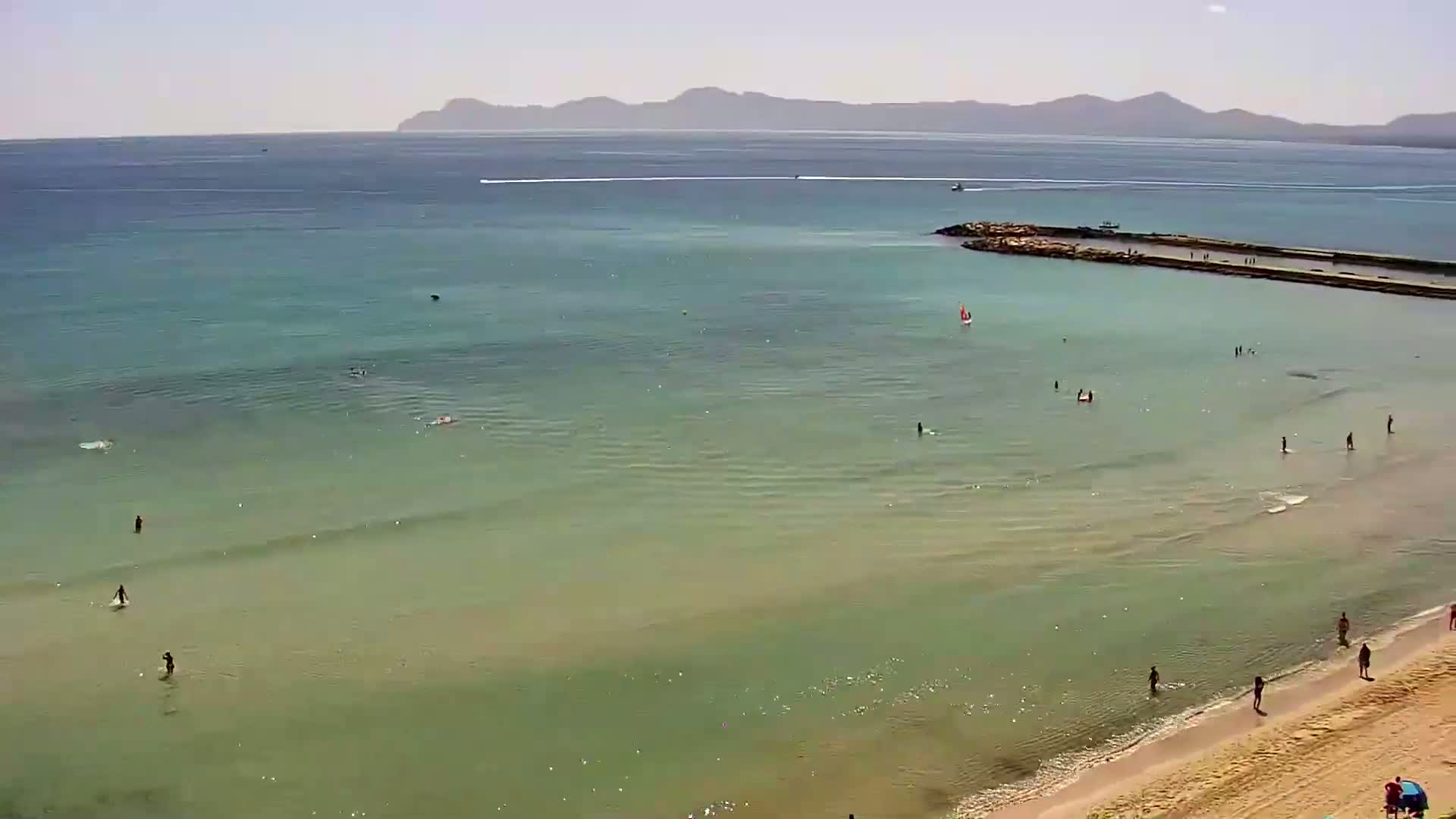 A sandy beach with calm, clear, light green water, a few people swimming and sunbathing, and distant mountains under a sunny sky.