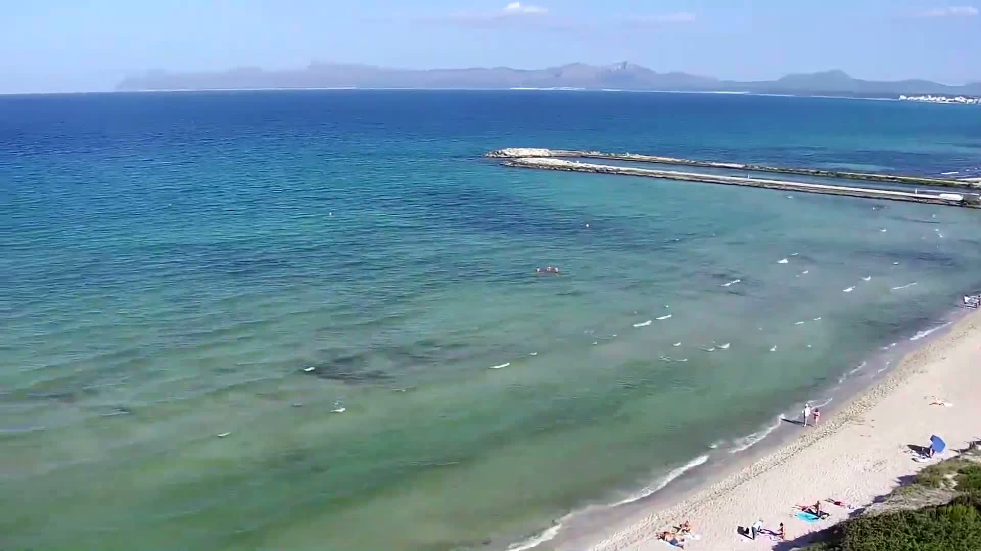 A sandy beach with several people on it slopes gently down to clear, light-green shallow water that deepens to a darker blue near a distant shoreline under a bright sunny sky.