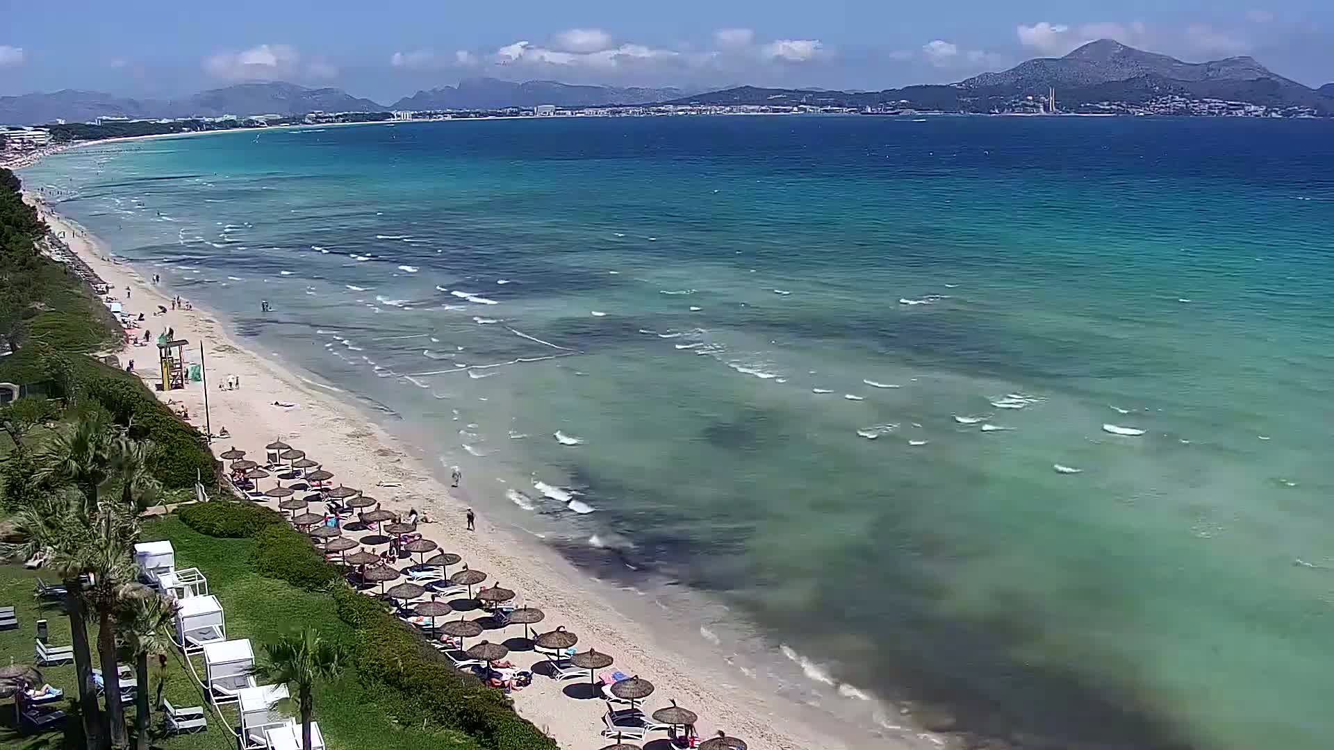 A sandy beach with numerous umbrellas and beachgoers stretches along a clear, turquoise ocean under a sunny sky, mountains visible in the distance.