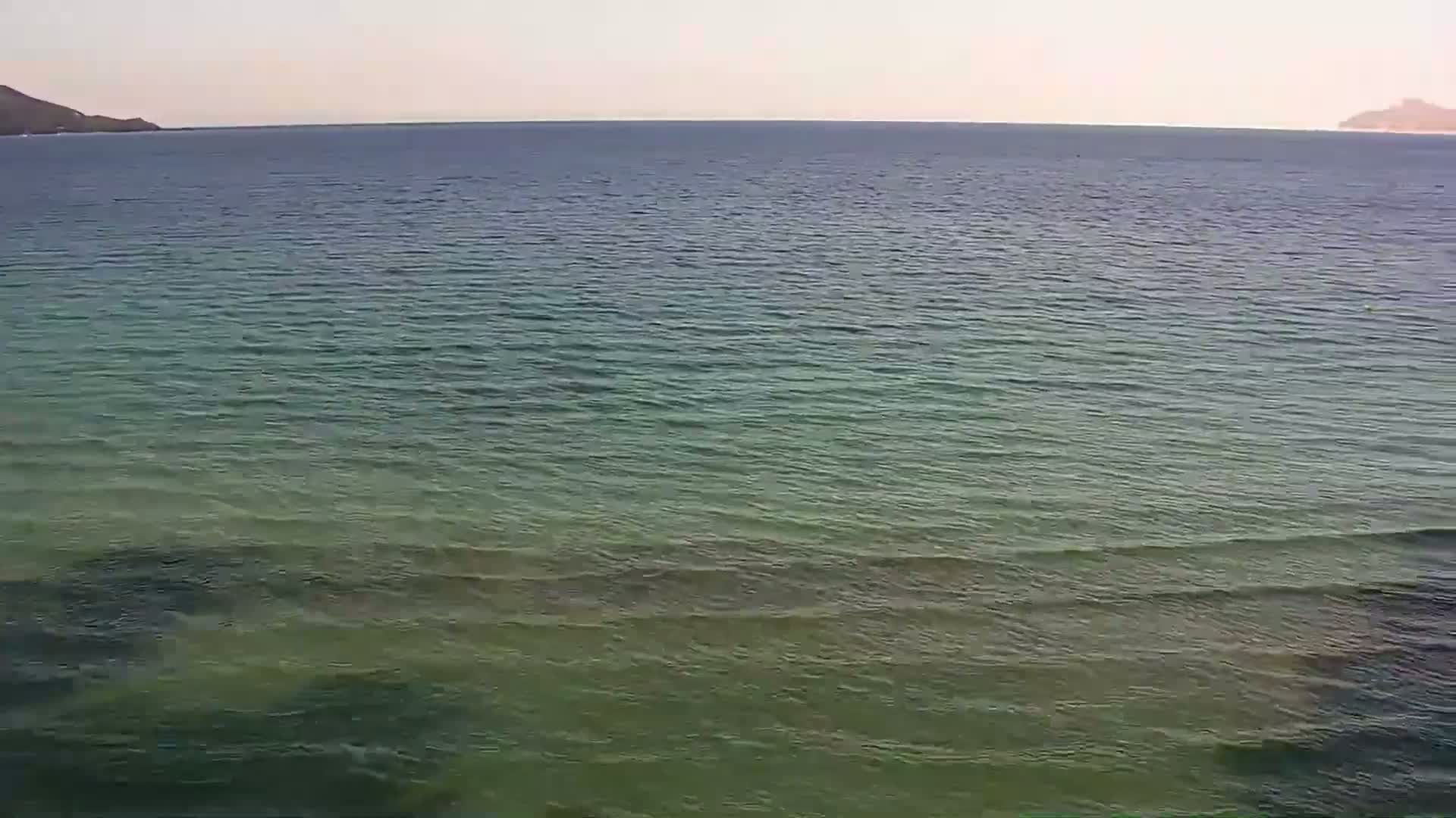 A calm, greenish-blue sea stretches to the horizon under a pale sky, with distant hills visible on either side.
