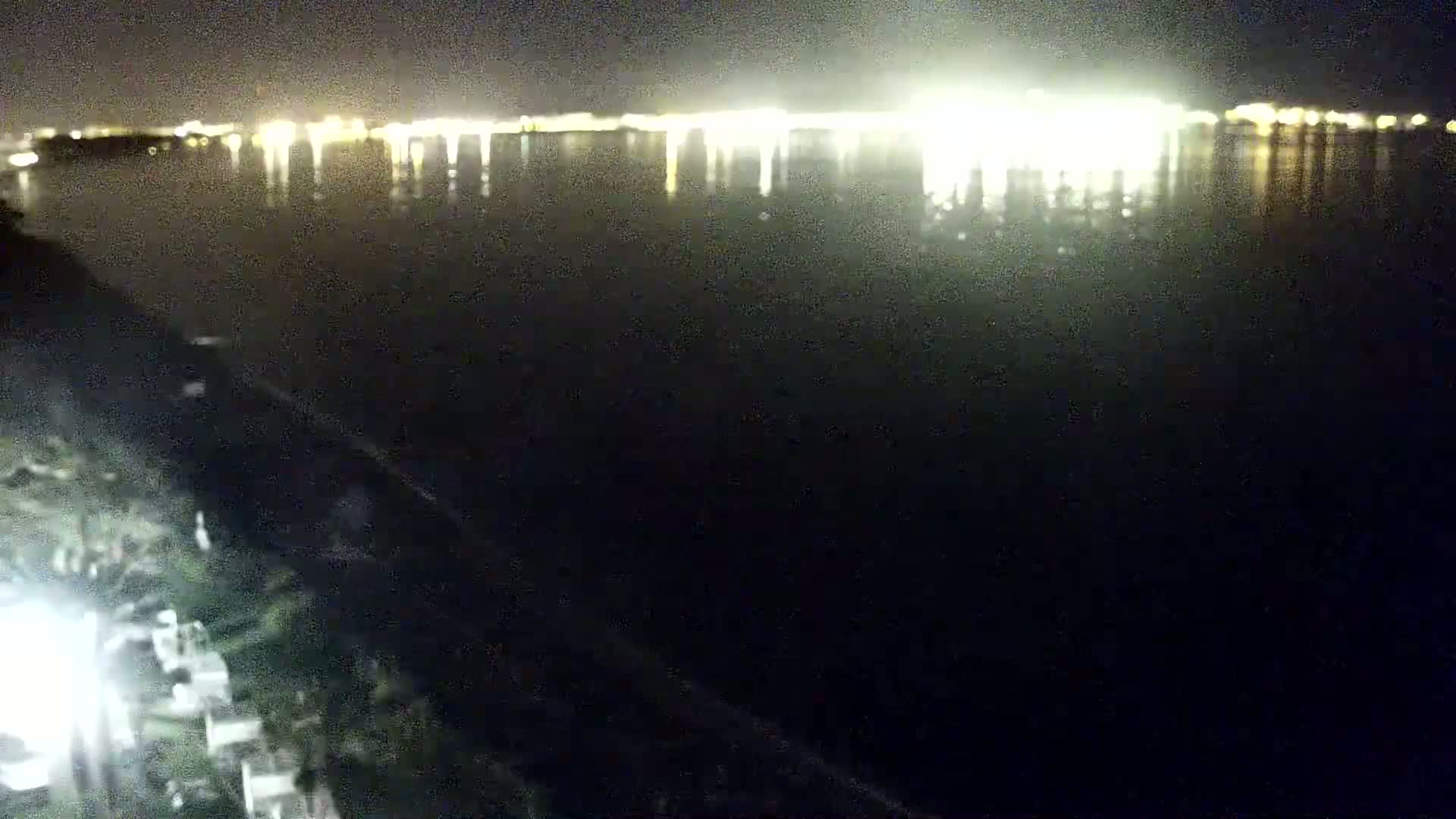A nighttime view shows a dark body of water reflecting the bright lights of a distant city across the water.