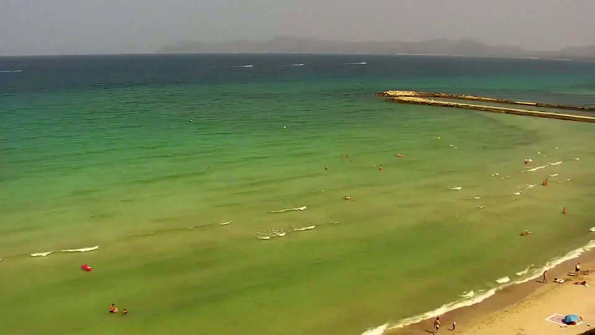 An aerial view shows a sandy beach with people swimming and sunbathing in clear, shallow, green-tinged water under a hazy sky.