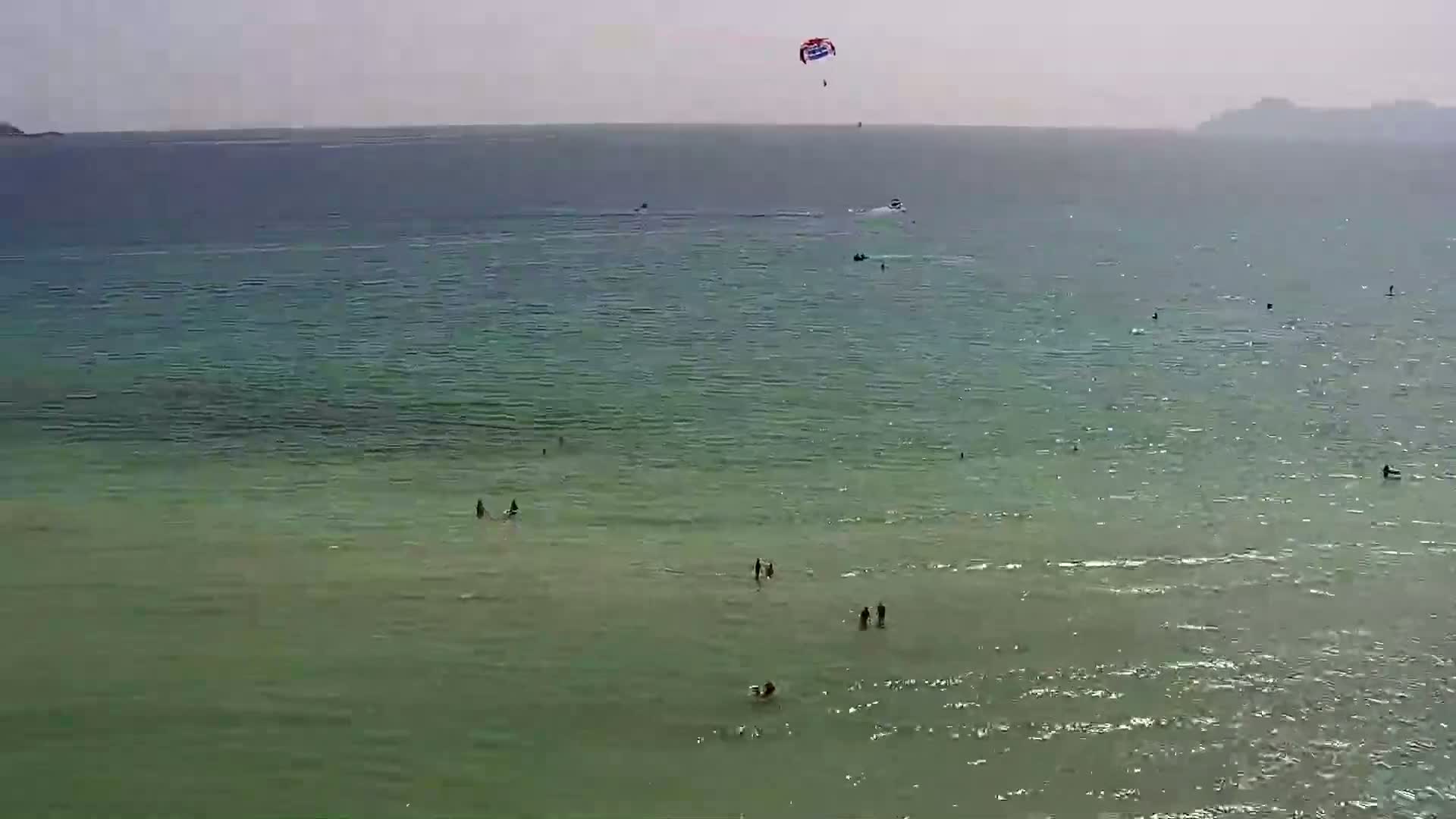 A parasail is in the air over a calm, light-green ocean where several people are swimming and a boat is visible in the distance.