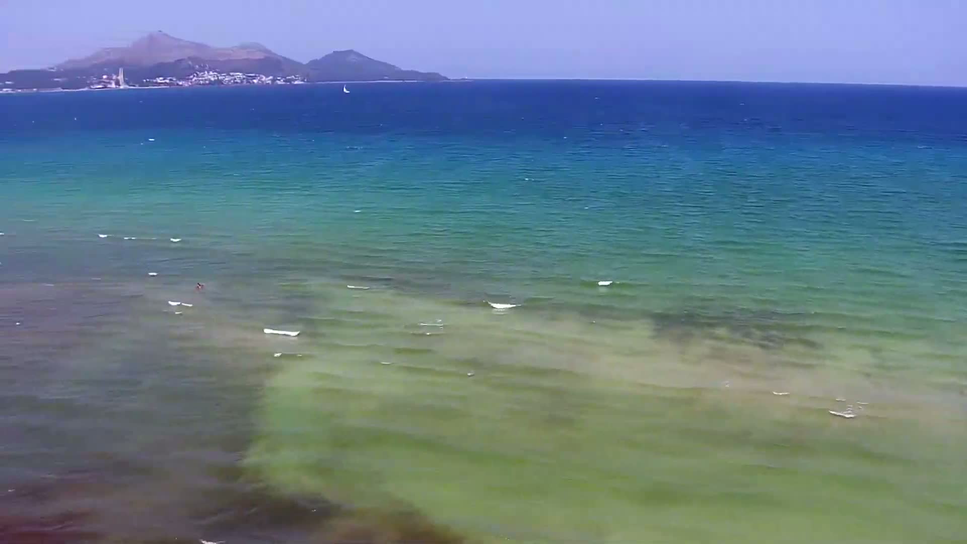 A calm, clear ocean with varying shades of blue and green, shallows near the shore, and a distant mountain range visible on the horizon.