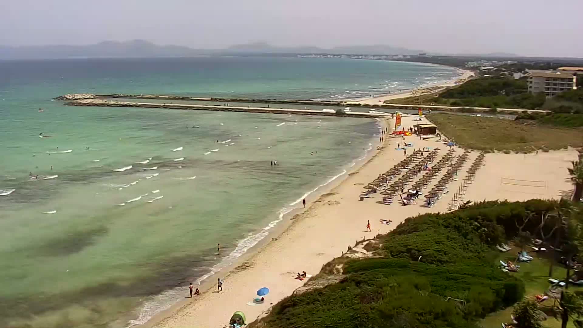 A sandy beach with calm, light-green water, numerous beach umbrellas, and a few people swimming and sunbathing is visible under a mostly clear sky.