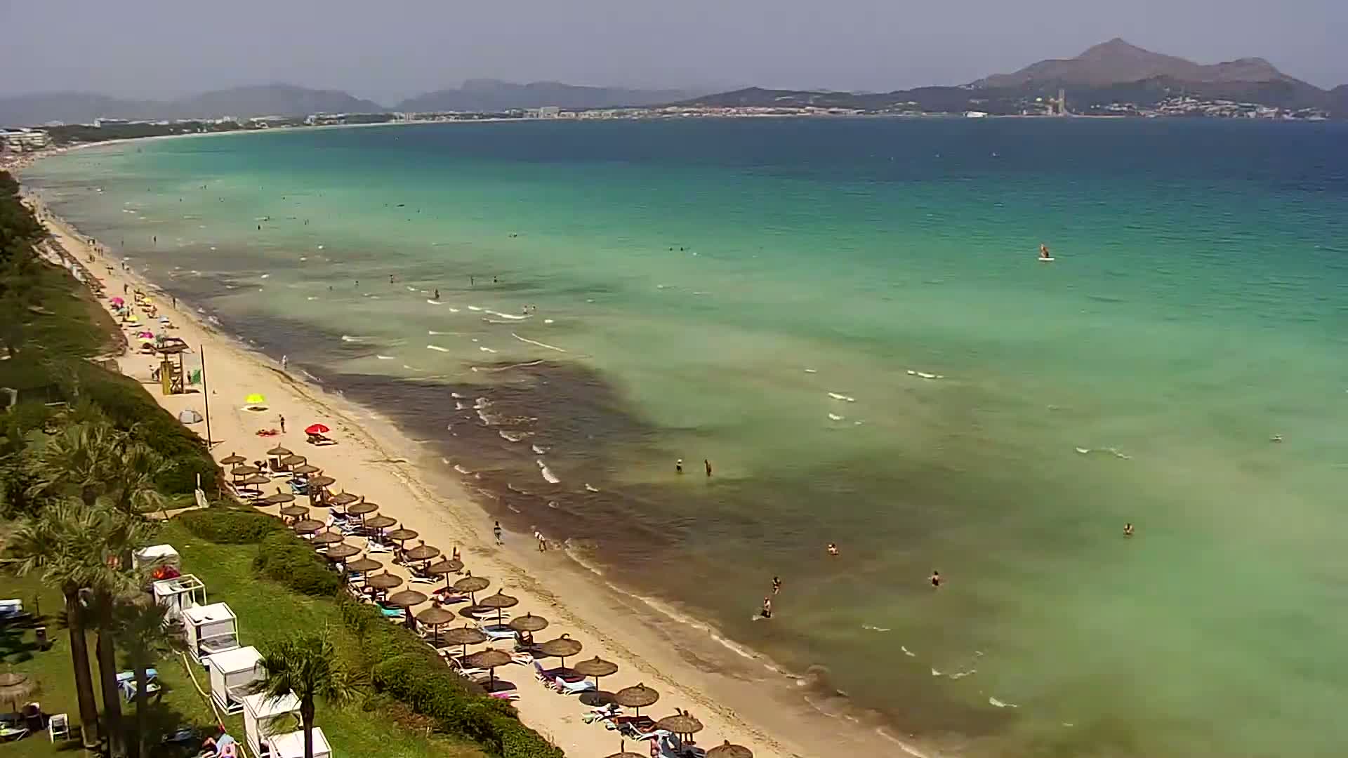 A sandy beach with turquoise water and numerous umbrellas lines the shore under a sunny sky, with a few people swimming and sunbathing.