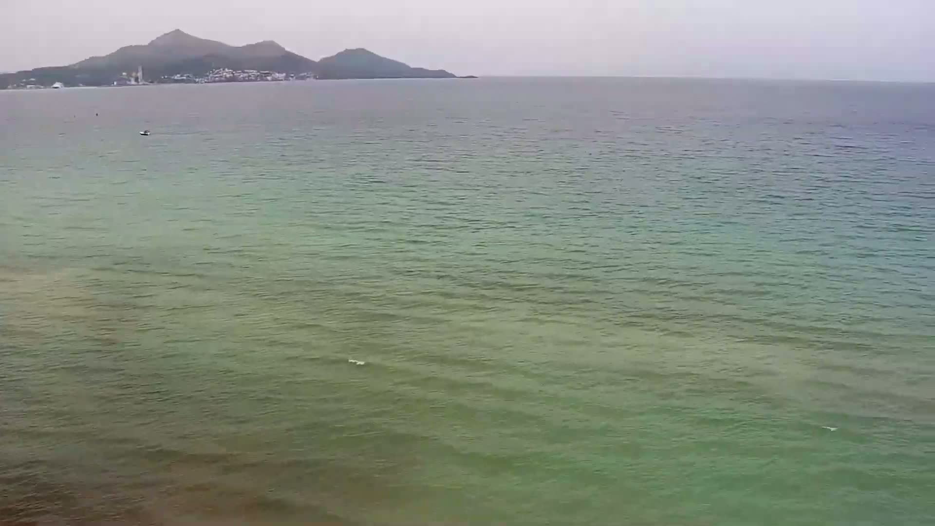 A calm, greenish-grey sea stretches to a distant, hazy mountain range under an overcast sky.