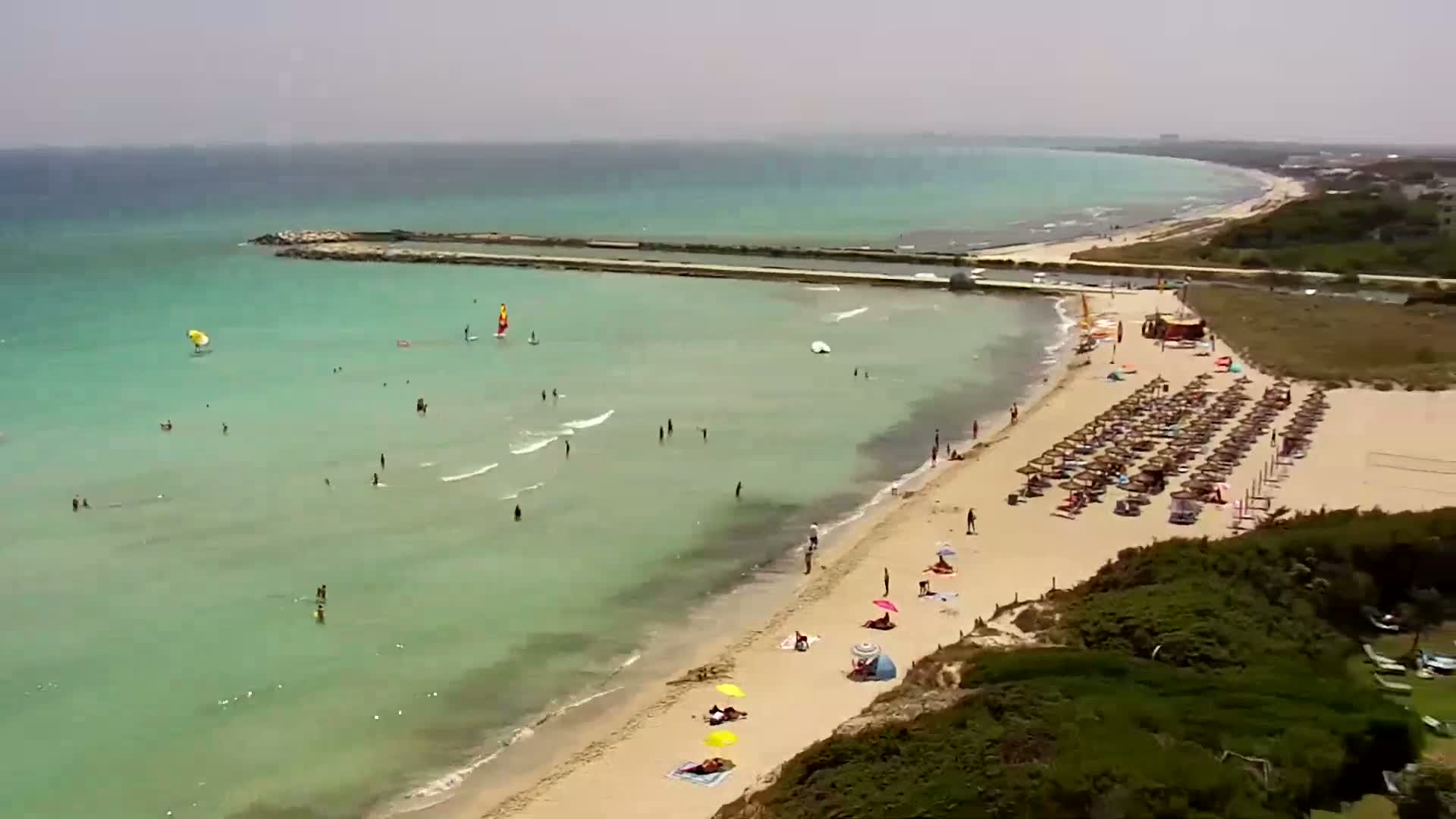 A sandy beach with many people swimming, sunbathing under umbrellas, and windsurfing in clear, shallow turquoise water on a sunny day.
