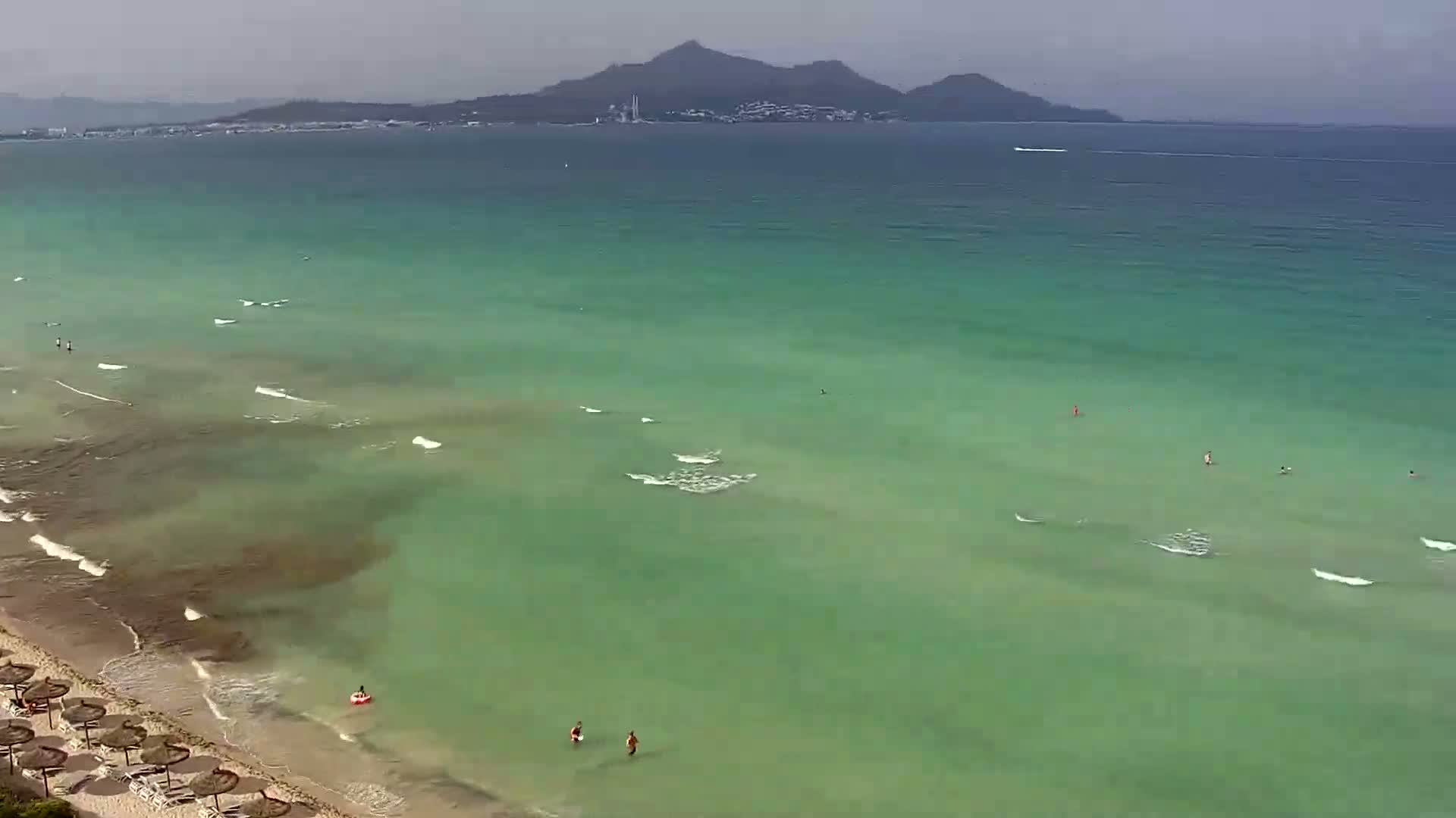 A mostly cloudy day shows a beach with a few people swimming in clear, teal water, and a distant mountain range visible on the horizon.