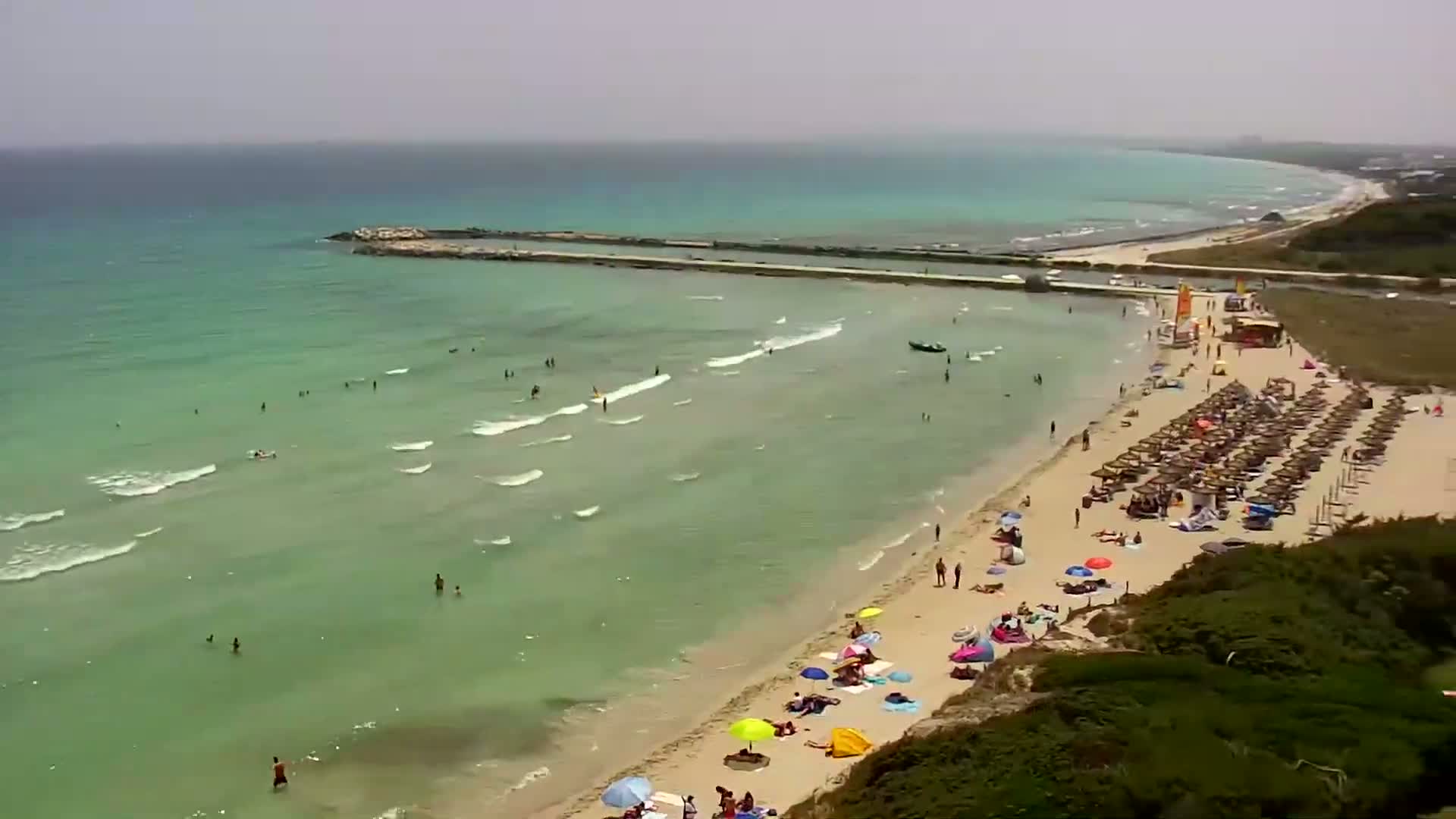 An aerial view shows a sandy beach with many people swimming and sunbathing under umbrellas on a sunny day with light waves in a calm, teal-colored sea.