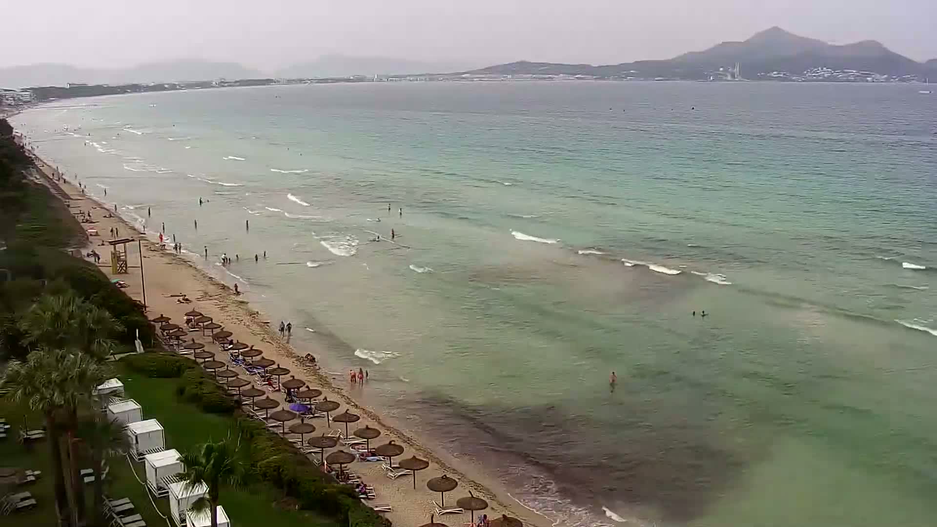 A sandy beach with light-colored umbrellas and people swimming in a calm, light-green sea under an overcast sky.