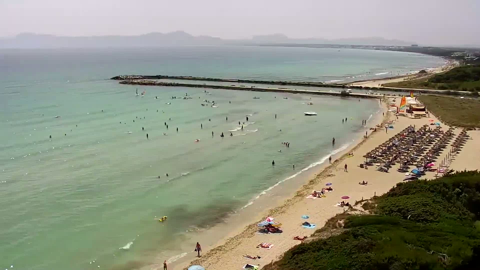 A sandy beach with many people swimming, sunbathing, and windsurfing in shallow, light-green water under a hazy sky.