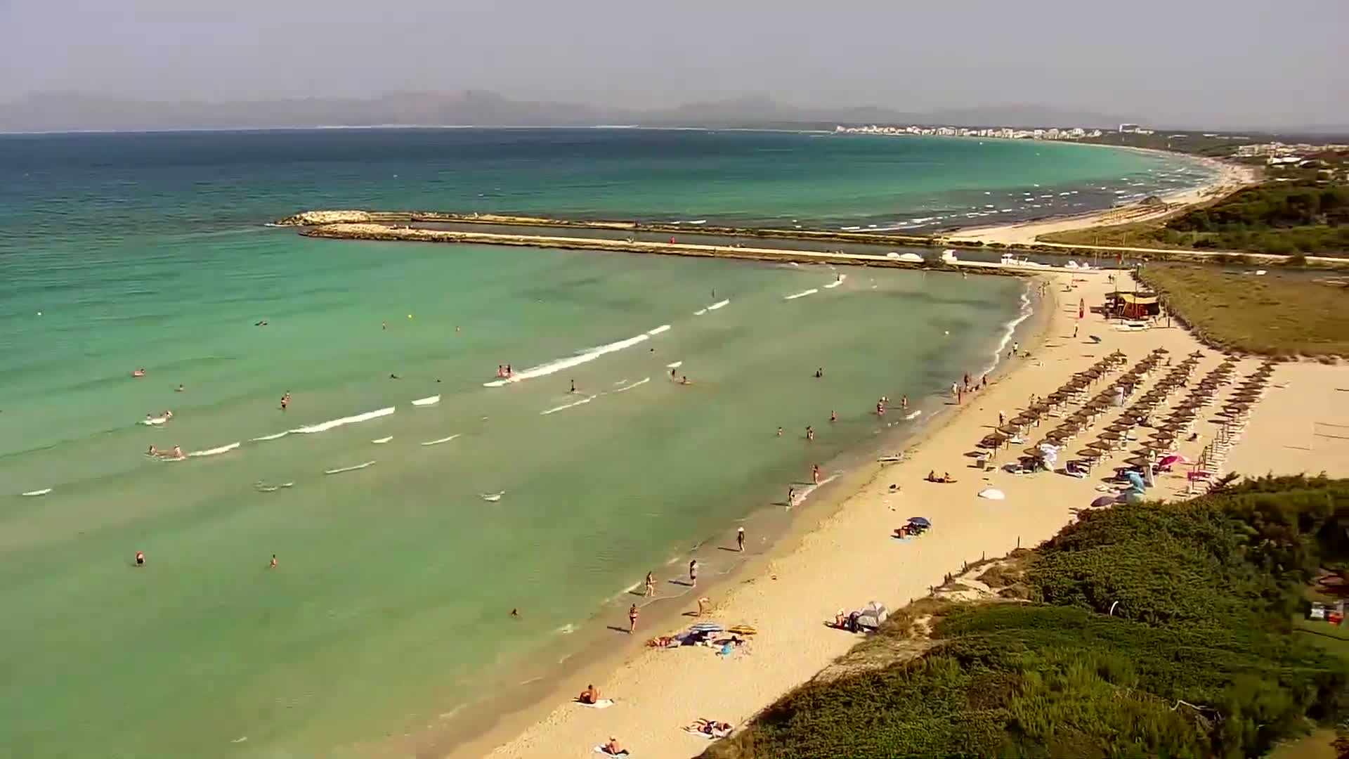 An aerial view shows a sandy beach with many people swimming and sunbathing under a clear, sunny sky, with calm, shallow, teal-colored water lapping the shore.