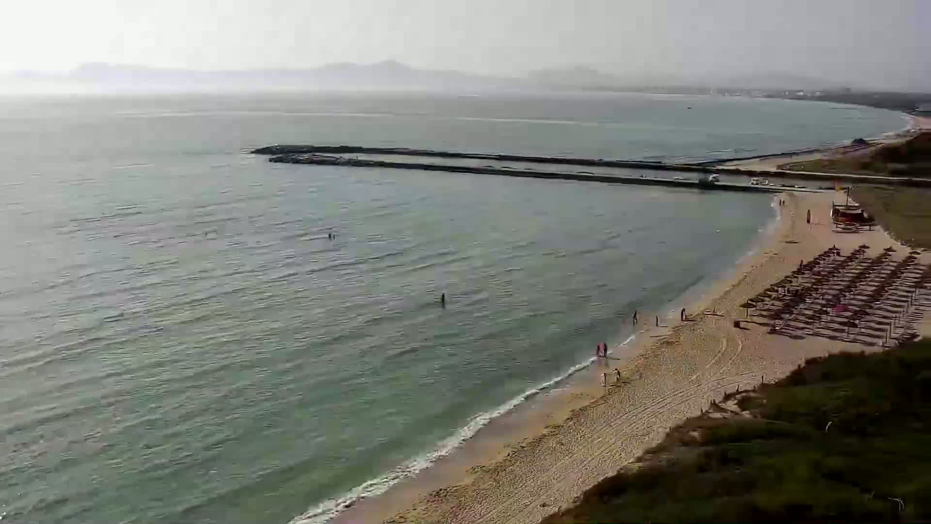A sandy beach with calm, light-green water and a hazy, distant coastline under a mostly clear sky.