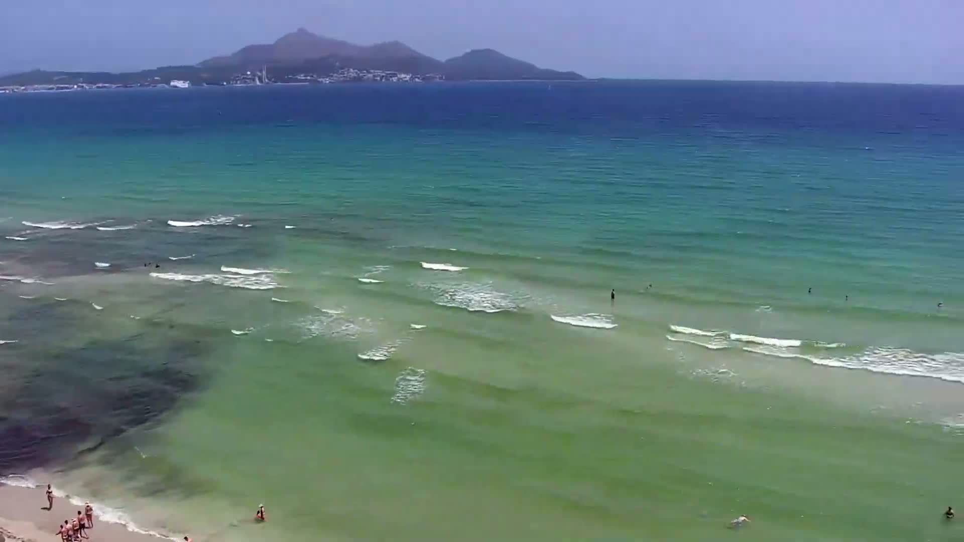 A sandy beach with people on it slopes down to a calm, shallow, green-tinged ocean with small waves under a clear, sunny sky, and a distant mountain range is visible on the horizon.