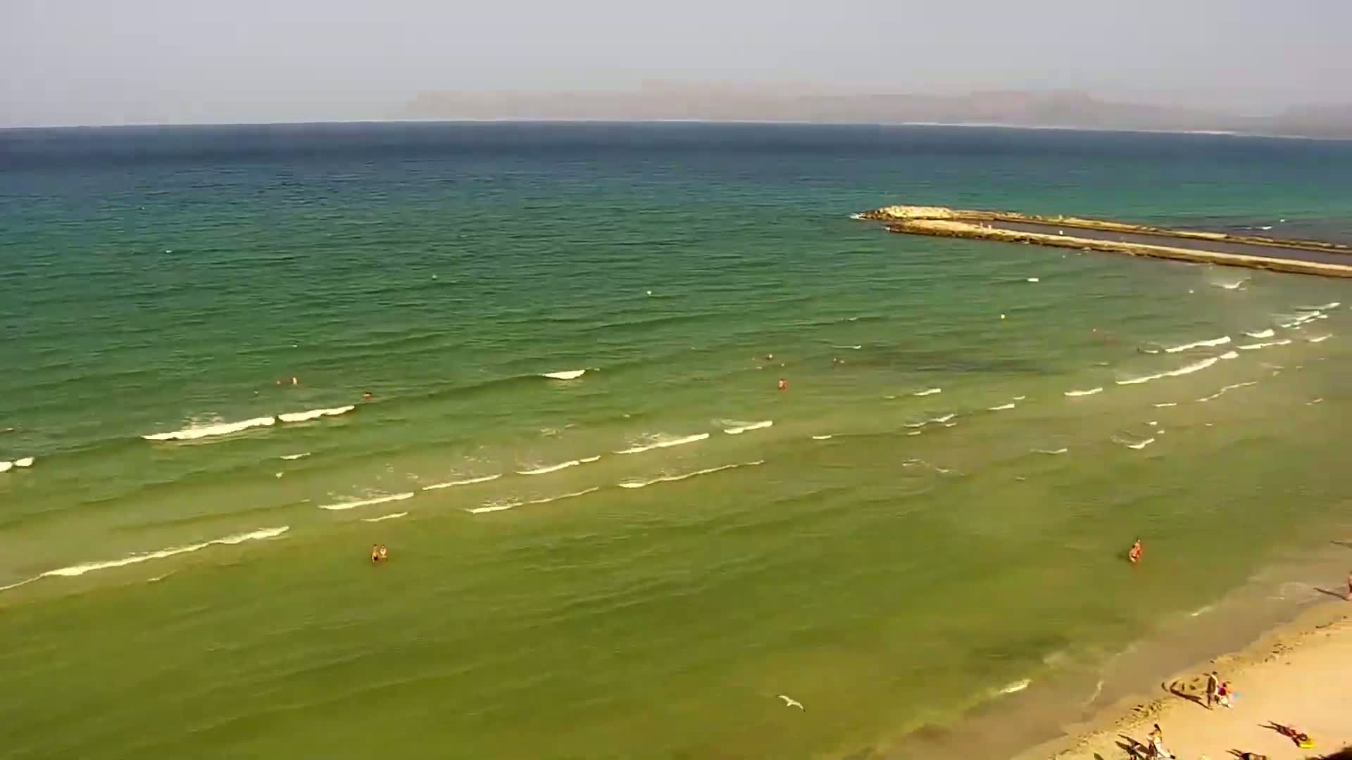 A sandy beach with a few people on it slopes down to a calm, green-tinged ocean with small waves under a clear sky.