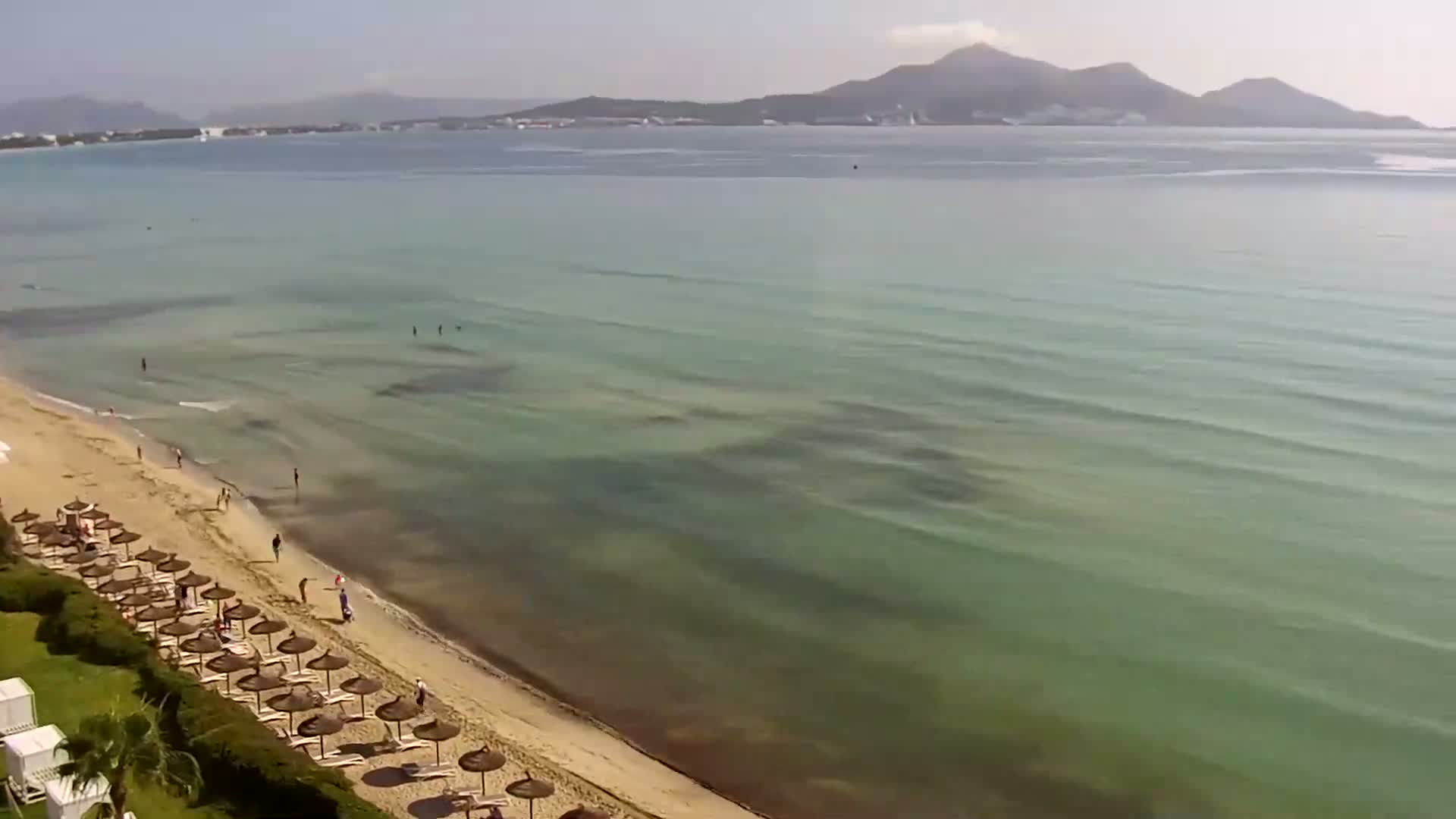 A sandy beach with numerous straw umbrellas and a few people is next to a calm, clear, green-tinged ocean under a mostly sunny sky, with mountains visible in the distance.