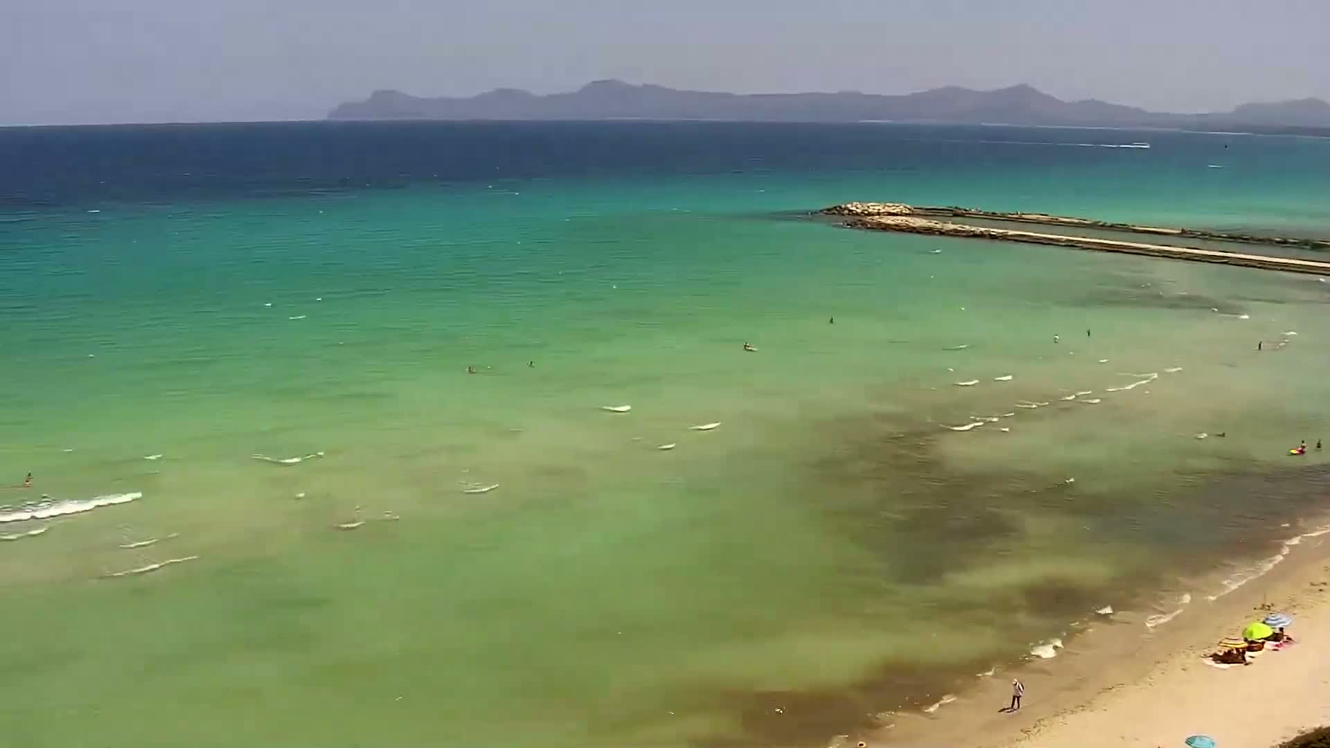 An aerial view shows a beach with a few people on the sand and in the shallow, clear, green-blue water under a mostly sunny sky, with distant mountains visible on the horizon.