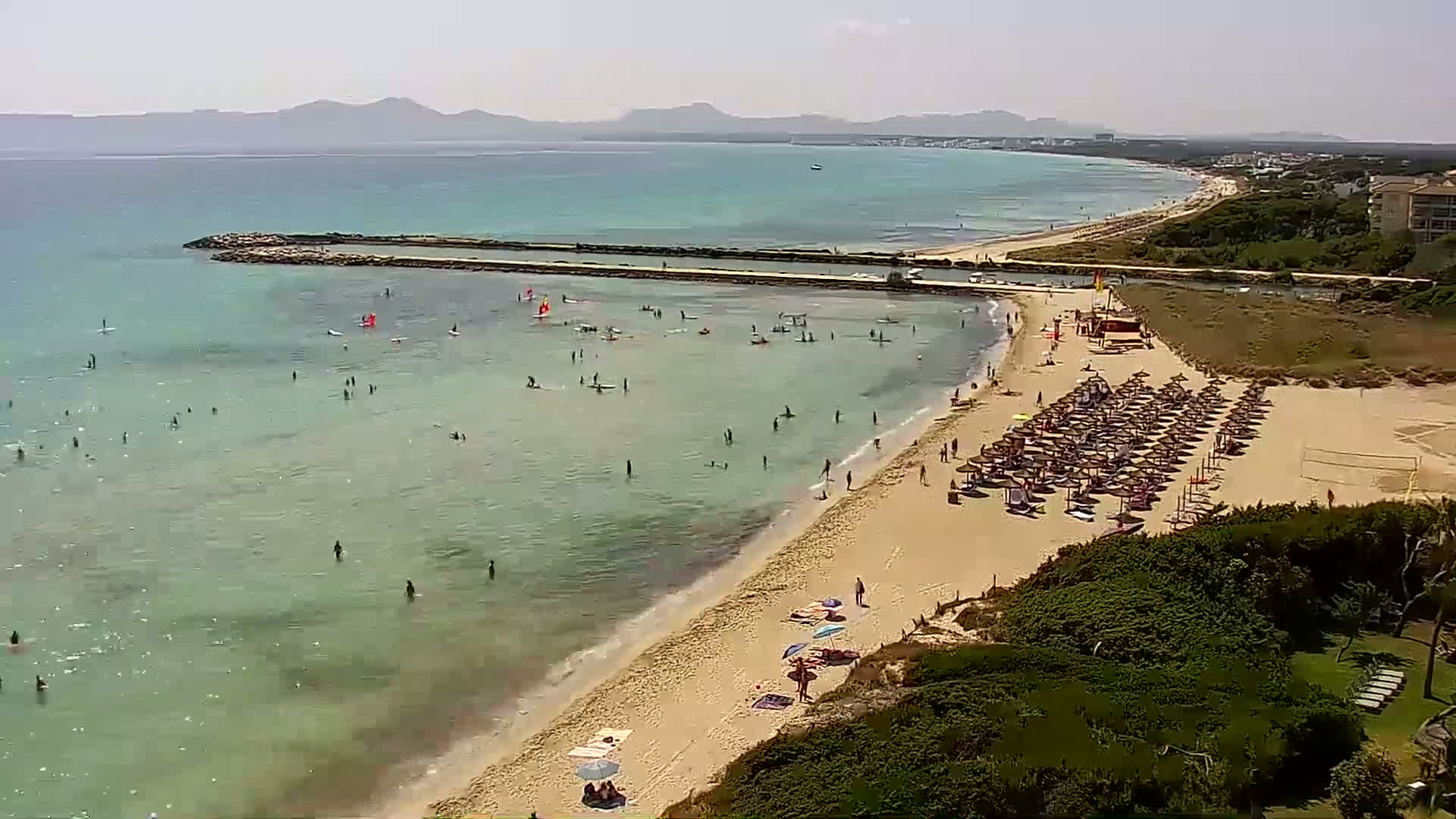 A sandy beach with numerous sunbathers and people swimming and windsurfing in clear, shallow water under a sunny sky, with distant mountains visible across a calm sea.