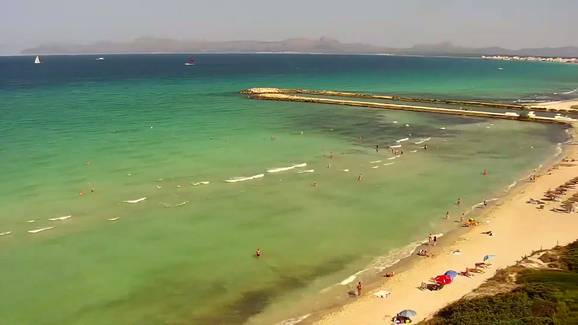 A sandy beach with many people swimming and sunbathing in clear, shallow, green water on a sunny day, with a distant coastline visible.
