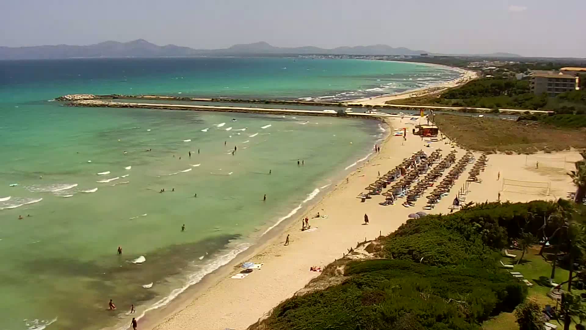 A sandy beach with turquoise water and many people swimming and sunbathing under a clear sunny sky, backed by low-lying vegetation and distant mountains.