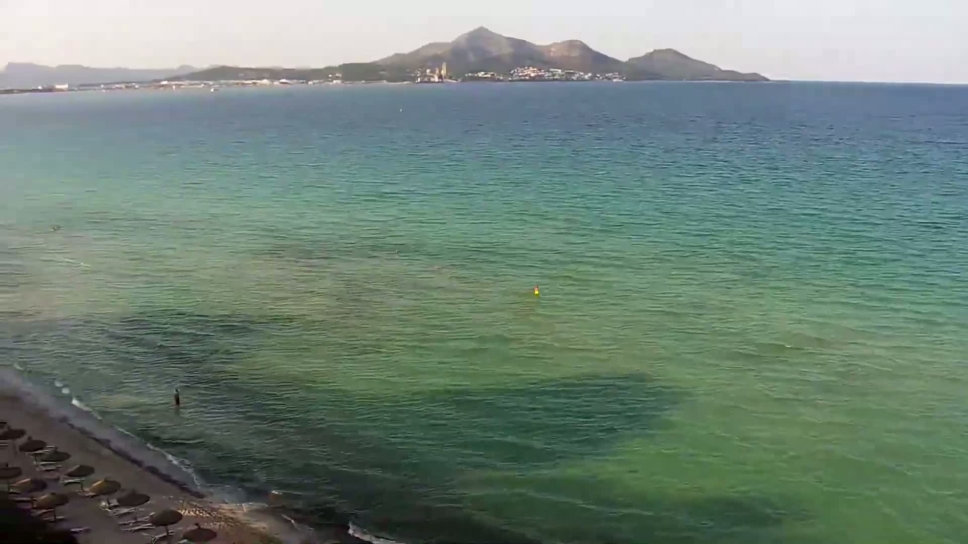 A sandy beach with a few umbrellas is next to a calm, clear, green-tinged ocean, with a distant mountain visible on the horizon under a hazy sky.