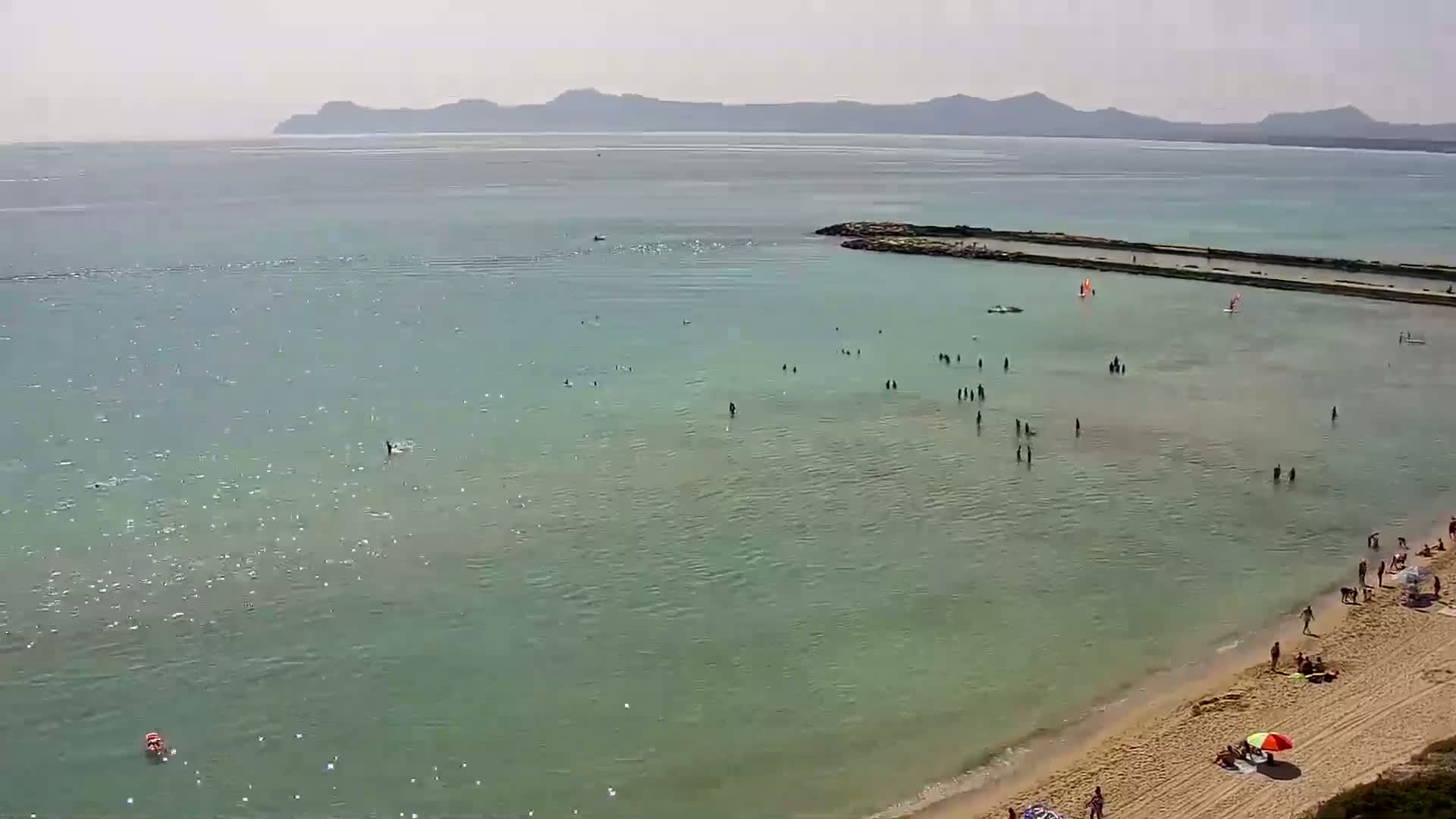 A sandy beach with many people swimming and sunbathing in calm, clear, light teal water under a sunny sky, with distant mountains visible in the background.