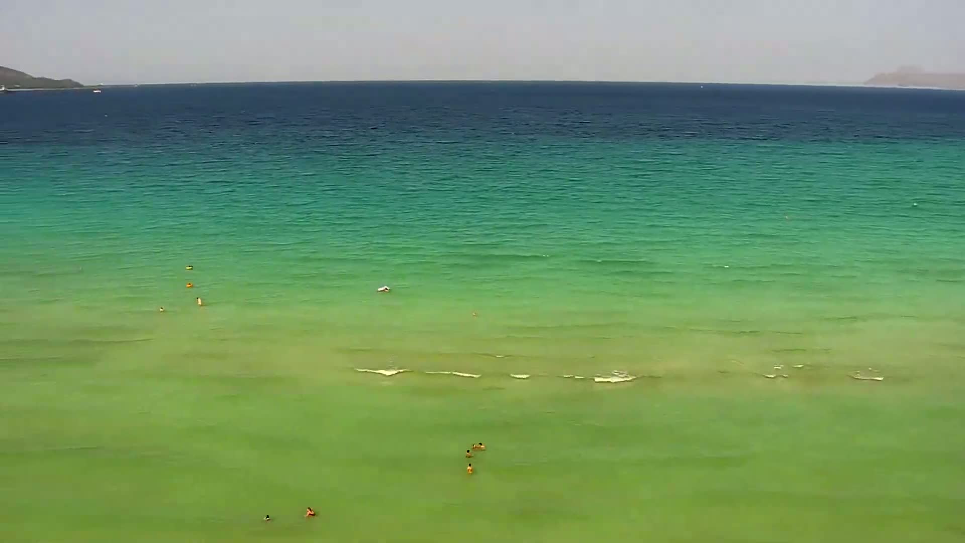 A calm, bright green ocean with several people swimming near the shore under a mostly cloudy sky.