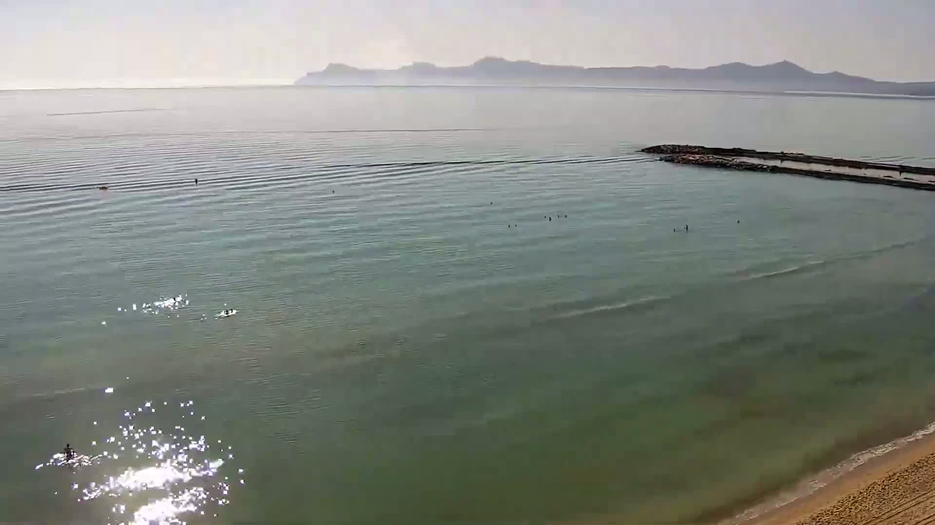 A calm, sunny day at a beach shows a few people swimming in clear, shallow water near a long jetty extending into a tranquil sea with distant mountains visible on the horizon.