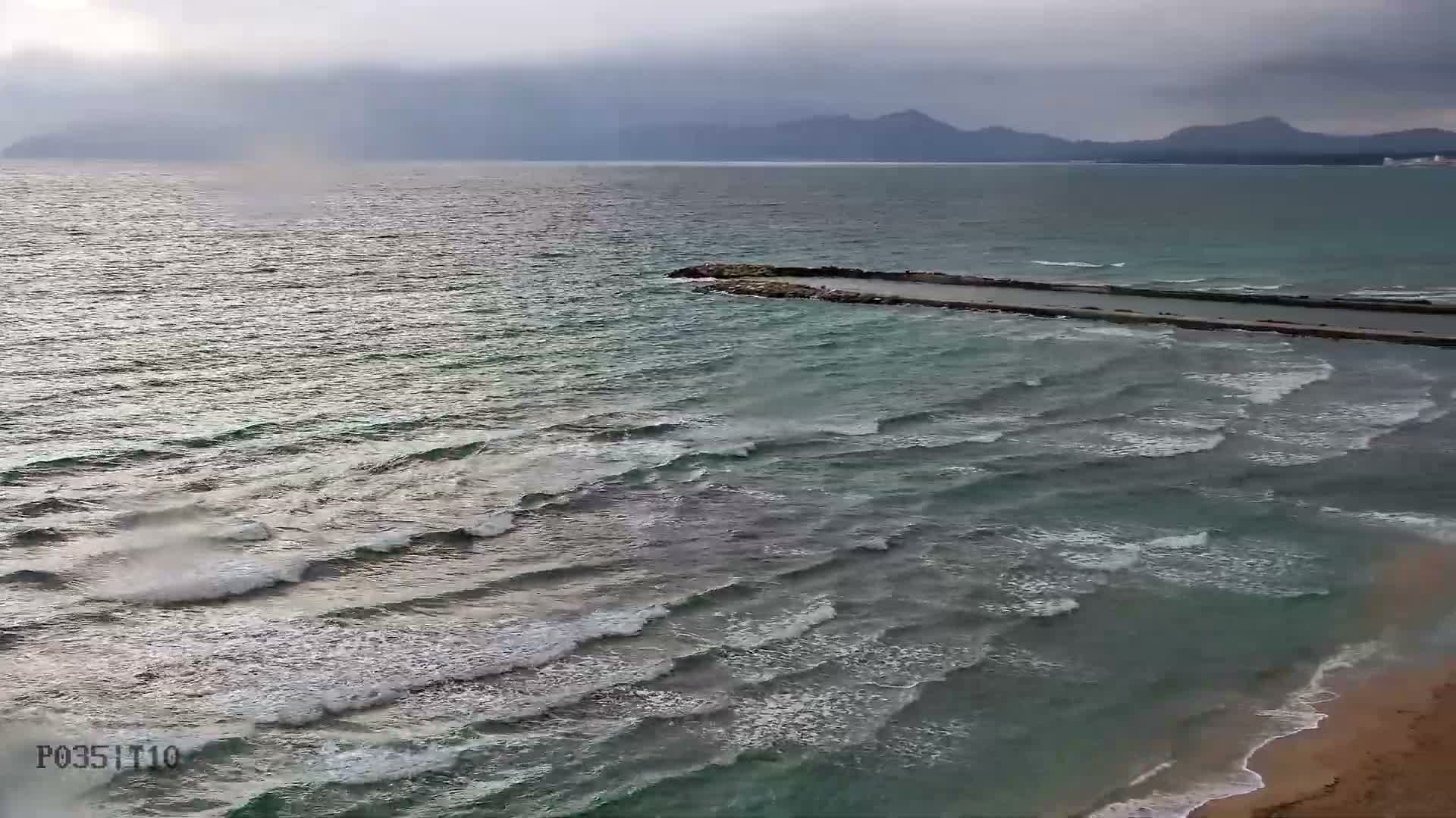 A choppy turquoise sea with white-capped waves breaks onto a sandy beach, bordered by a long concrete breakwater under an overcast sky with distant, hazy mountains.