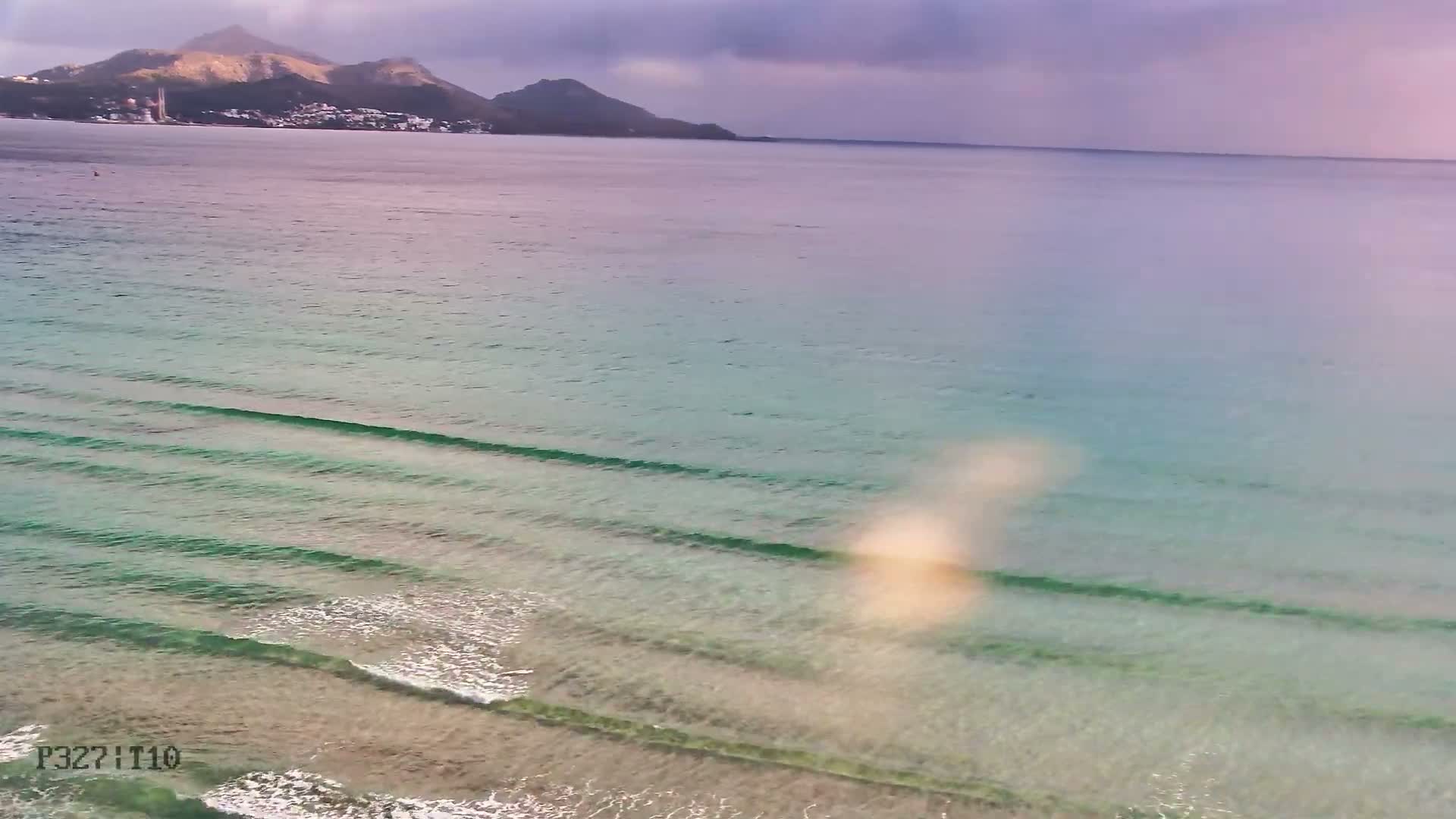 Gentle waves break on a clear, sandy beach leading to a calm ocean, with a mountainous coastline in the distance under an overcast, purplish sky.