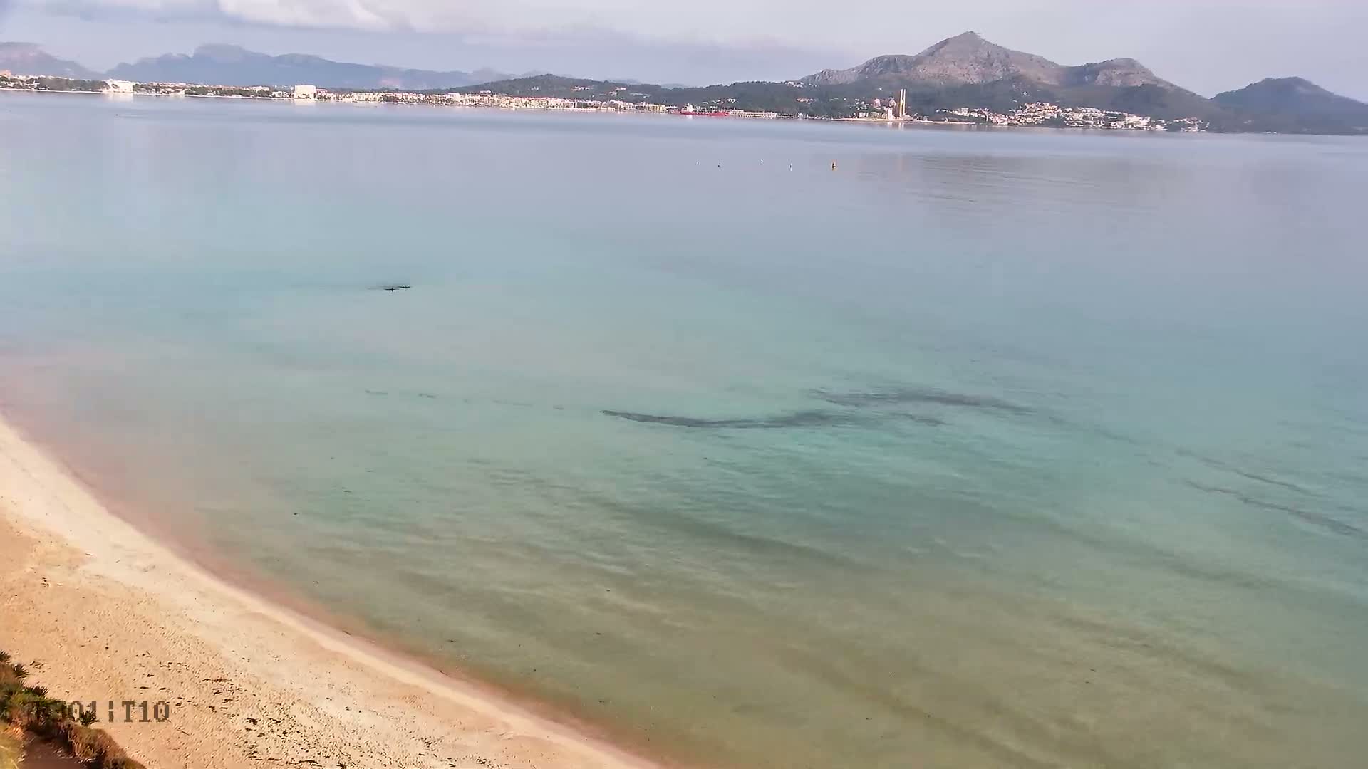 A tranquil, clear turquoise bay with a sandy beach in the foreground is visible under a partly cloudy sky, with a distant town and mountainous coastline across the water.