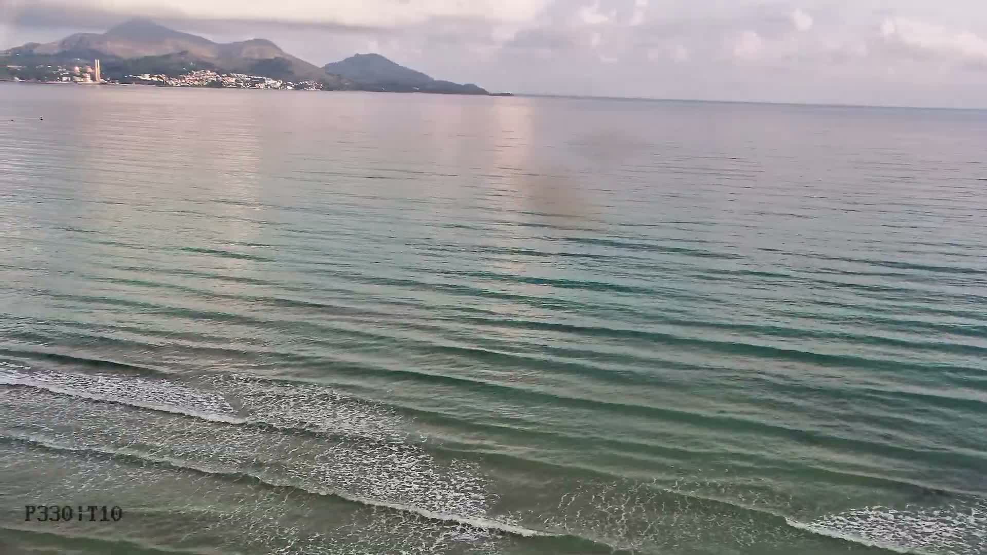 A tranquil, clear turquoise bay with a sandy beach in the foreground is visible under a partly cloudy sky, with a distant town and mountainous coastline across the water.