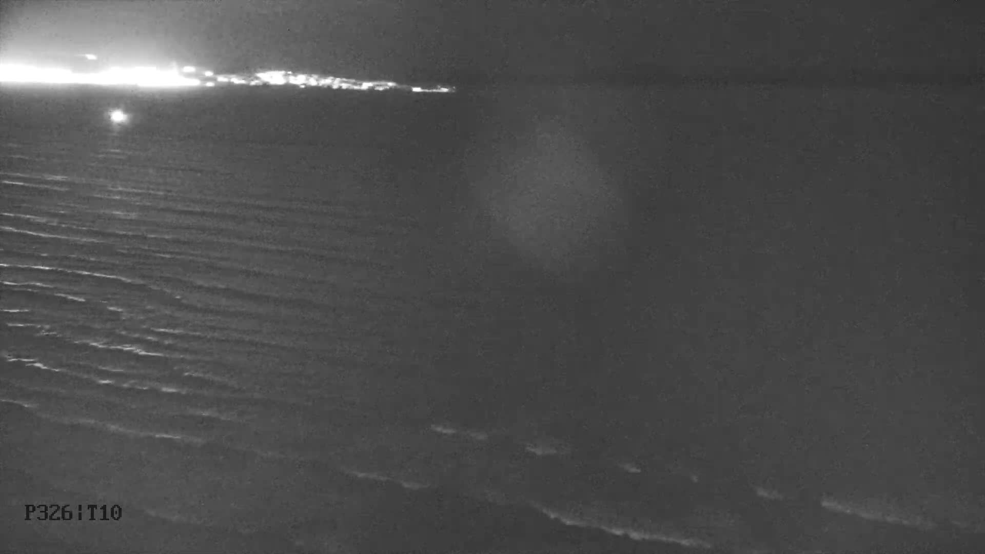 A tranquil, clear turquoise bay with a sandy beach in the foreground is visible under a partly cloudy sky, with a distant town and mountainous coastline across the water.